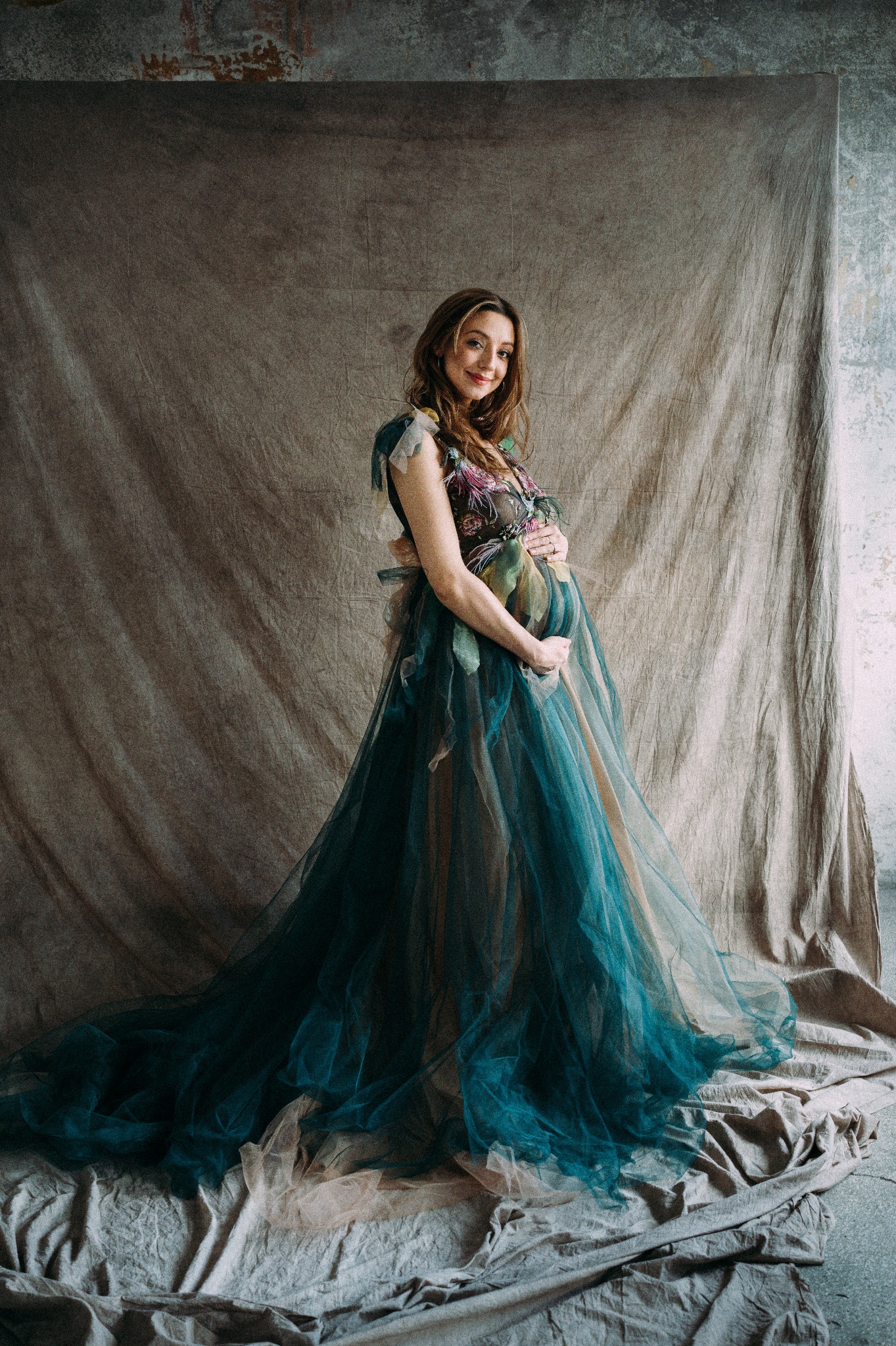 Kobieta w ciąży w długiej, tiulowej sukni z kwiatowymi aplikacjami, pozująca na tle teksturowanej tkaniny i ściany.