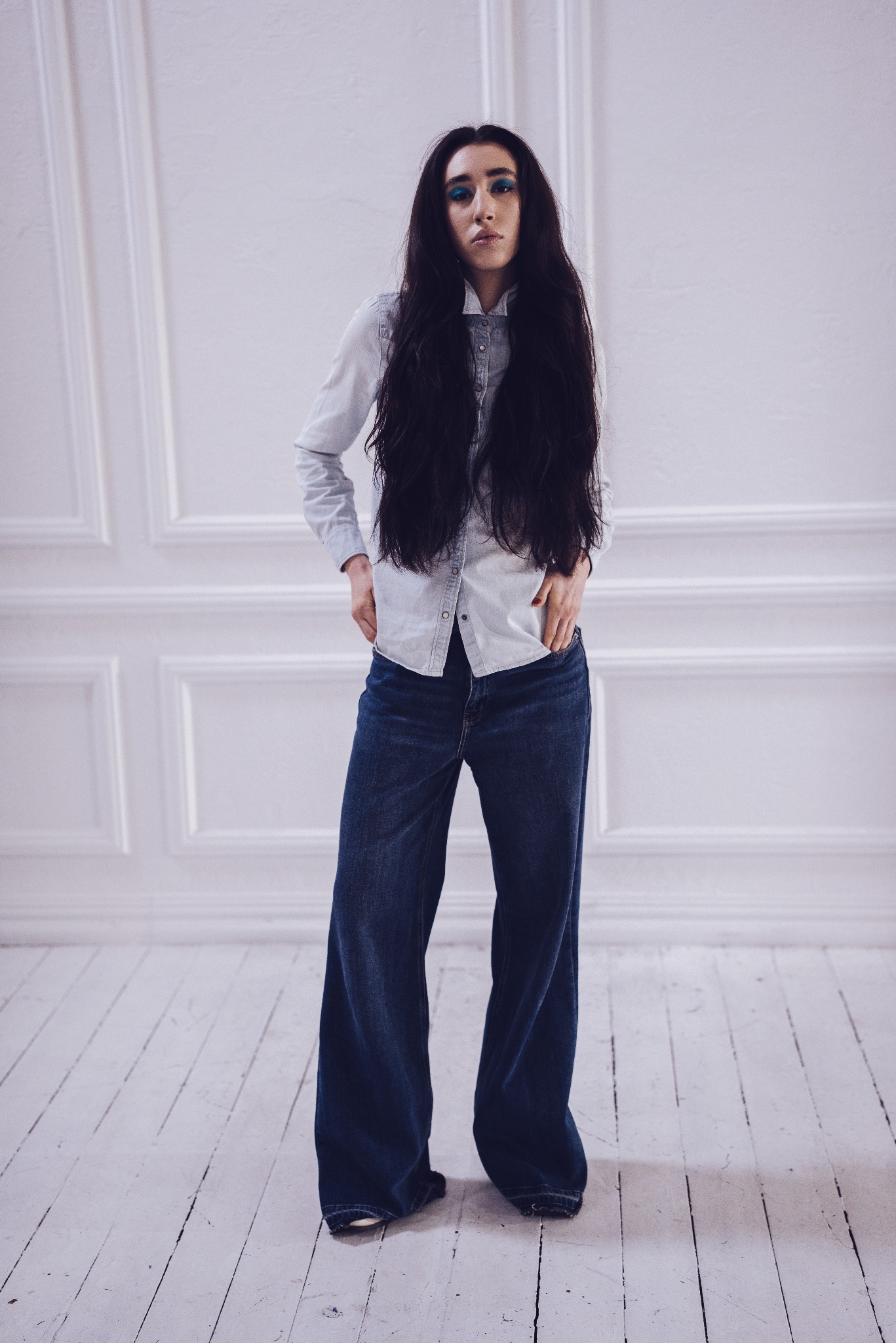 Kobieta w dżinsach z szerokimi nogawkami, jasnej koszuli i z intensywnym, niebieskim makijażem oczu, stojąca na tle białej ściany z dekoracyjnymi panelami.
