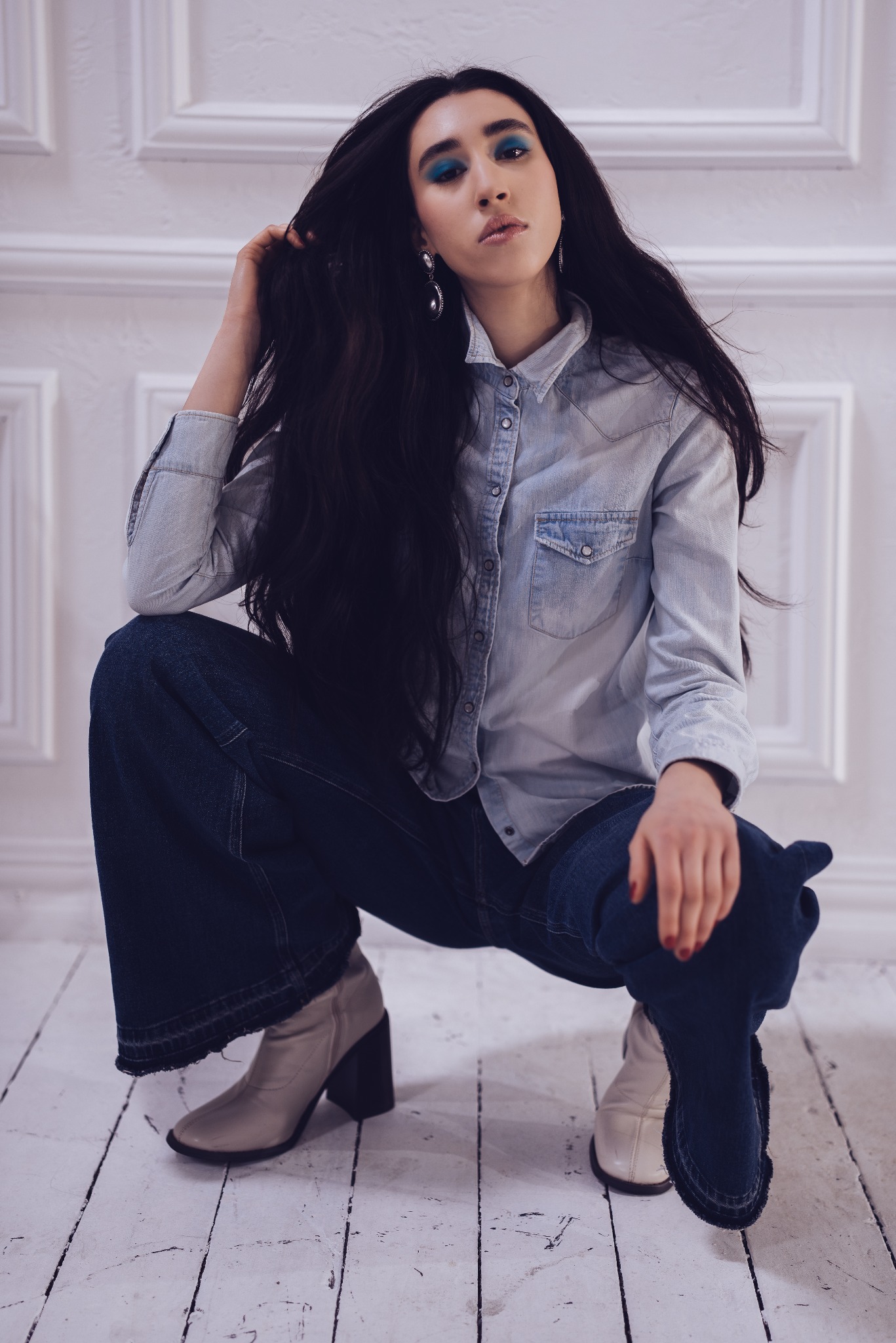 Kobieta w dżinsowej koszuli i szerokich spodniach, pozująca w przysiadzie na białej podłodze, z intensywnym niebieskim makijażem oczu i dużymi kolczykami.