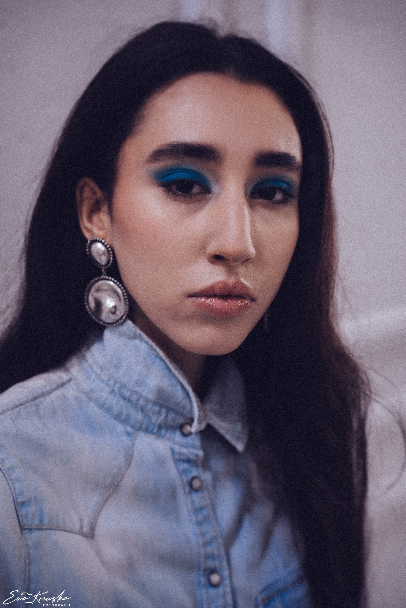 Portret kobiety z intensywnym niebieskim makijażem oczu, dużymi srebrnymi kolczykami i w dżinsowej koszuli, wykonany w stonowanej kolorystyce.