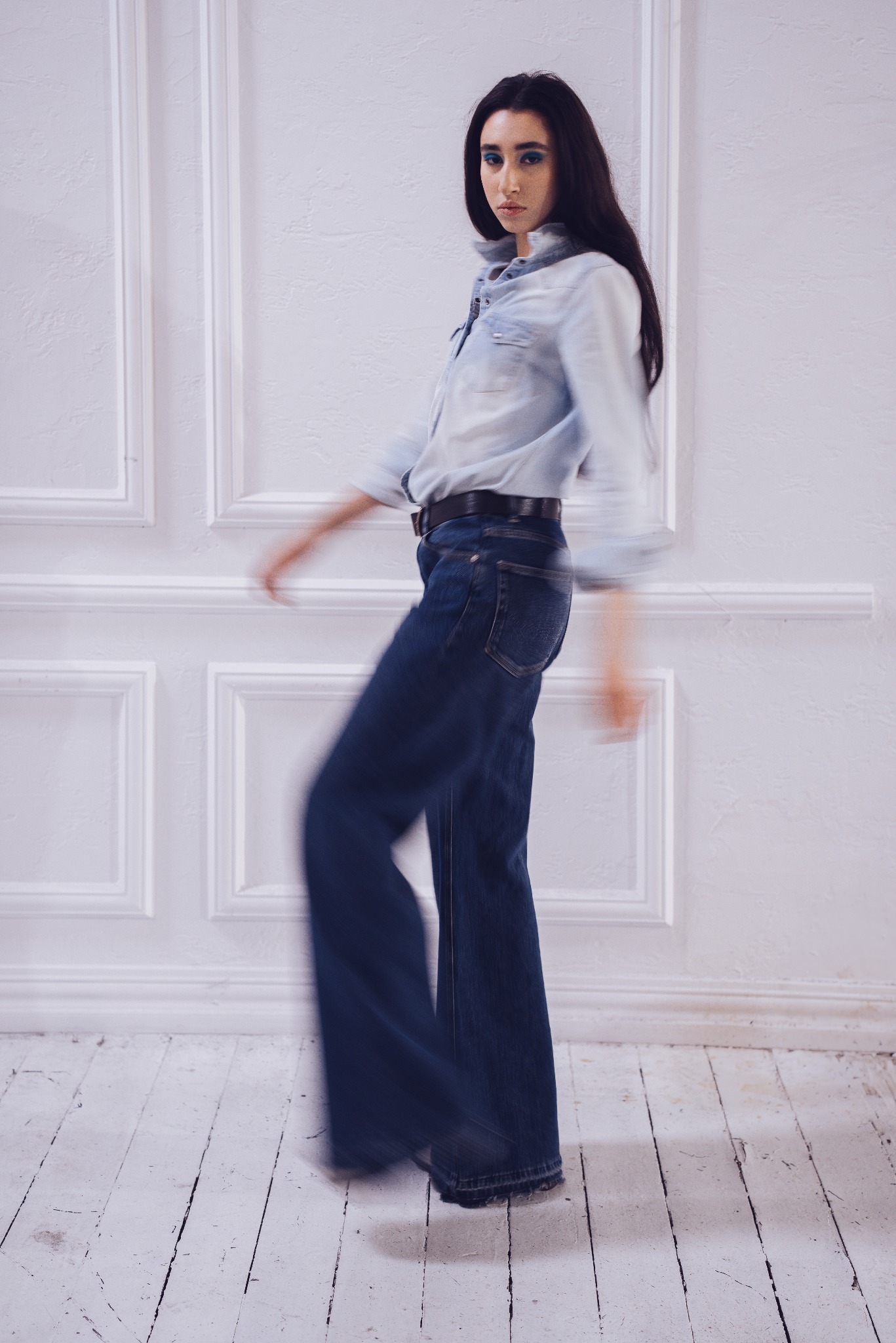 Kobieta w ruchu, ubrana w jeansową koszulę i szerokie spodnie, pozuje na tle białej ściany z dekoracyjnymi panelami.
