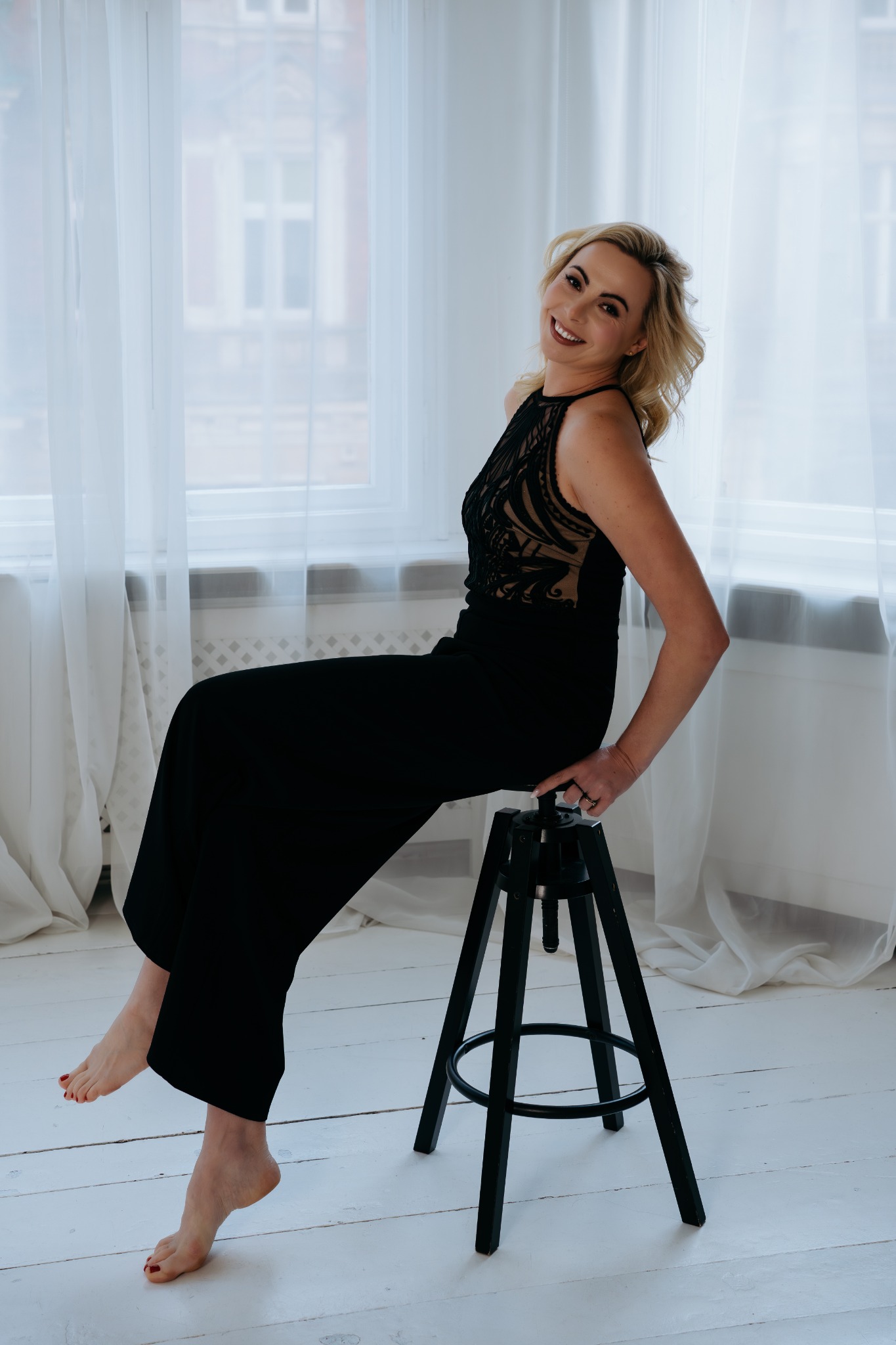 Uśmiechnięta blondynka w czarnym kombinezonie z koronkową górą, siedząca na czarnym stołku na tle okna z białymi zasłonami.