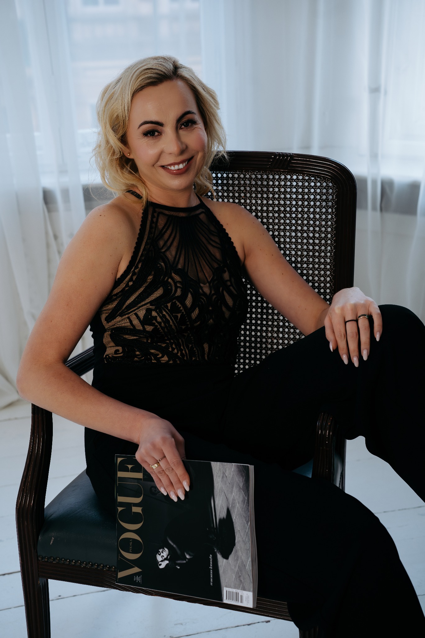 Portret uśmiechniętej blondynki w czarnym kombinezonie siedzącej w fotelu z magazynem Vogue w ręku, na tle jasnych zasłon.