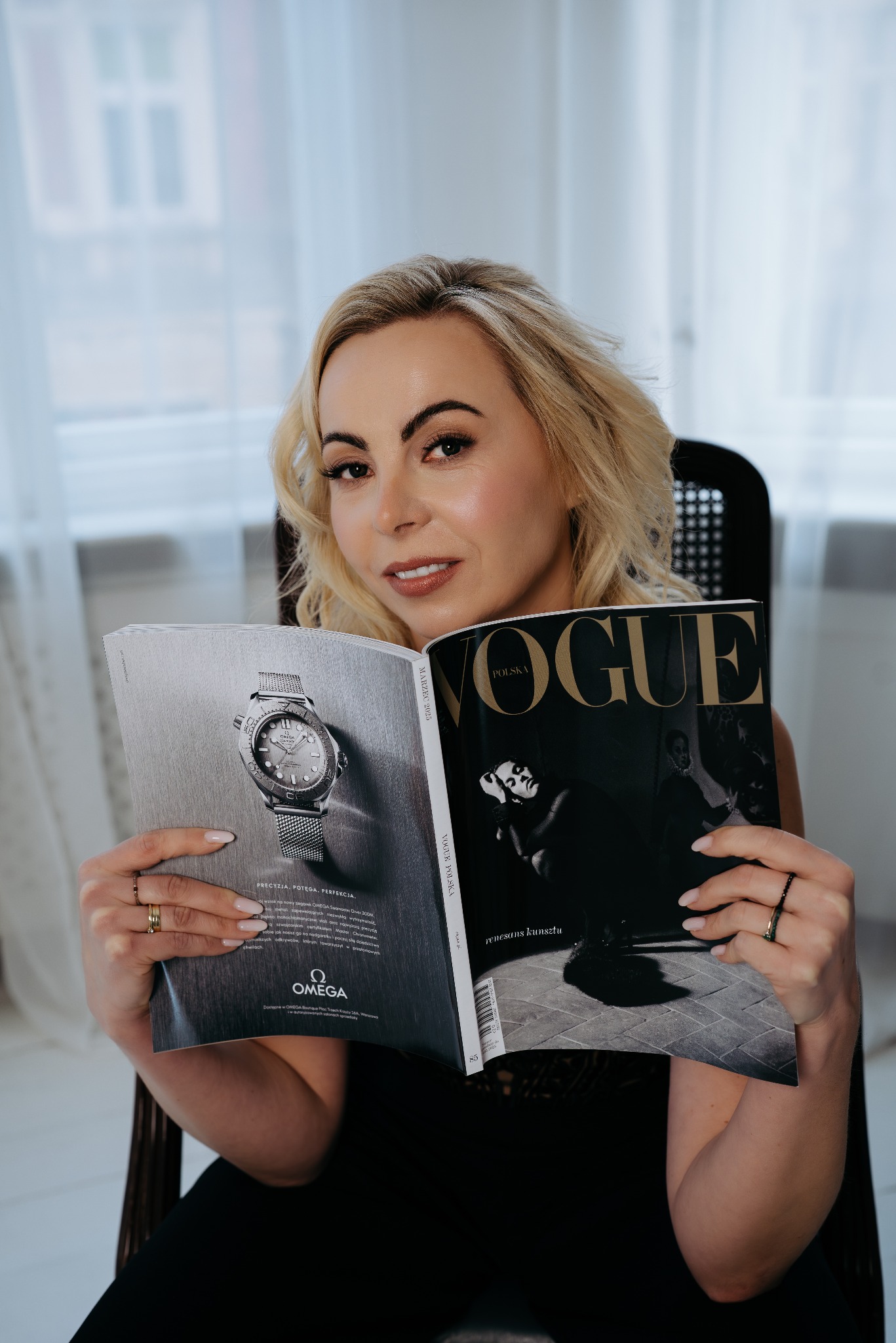 Kobieta z blond włosami, siedząca w fotelu, czyta magazyn Vogue Polska z reklamą zegarka Omega na okładce, uśmiechając się do aparatu.