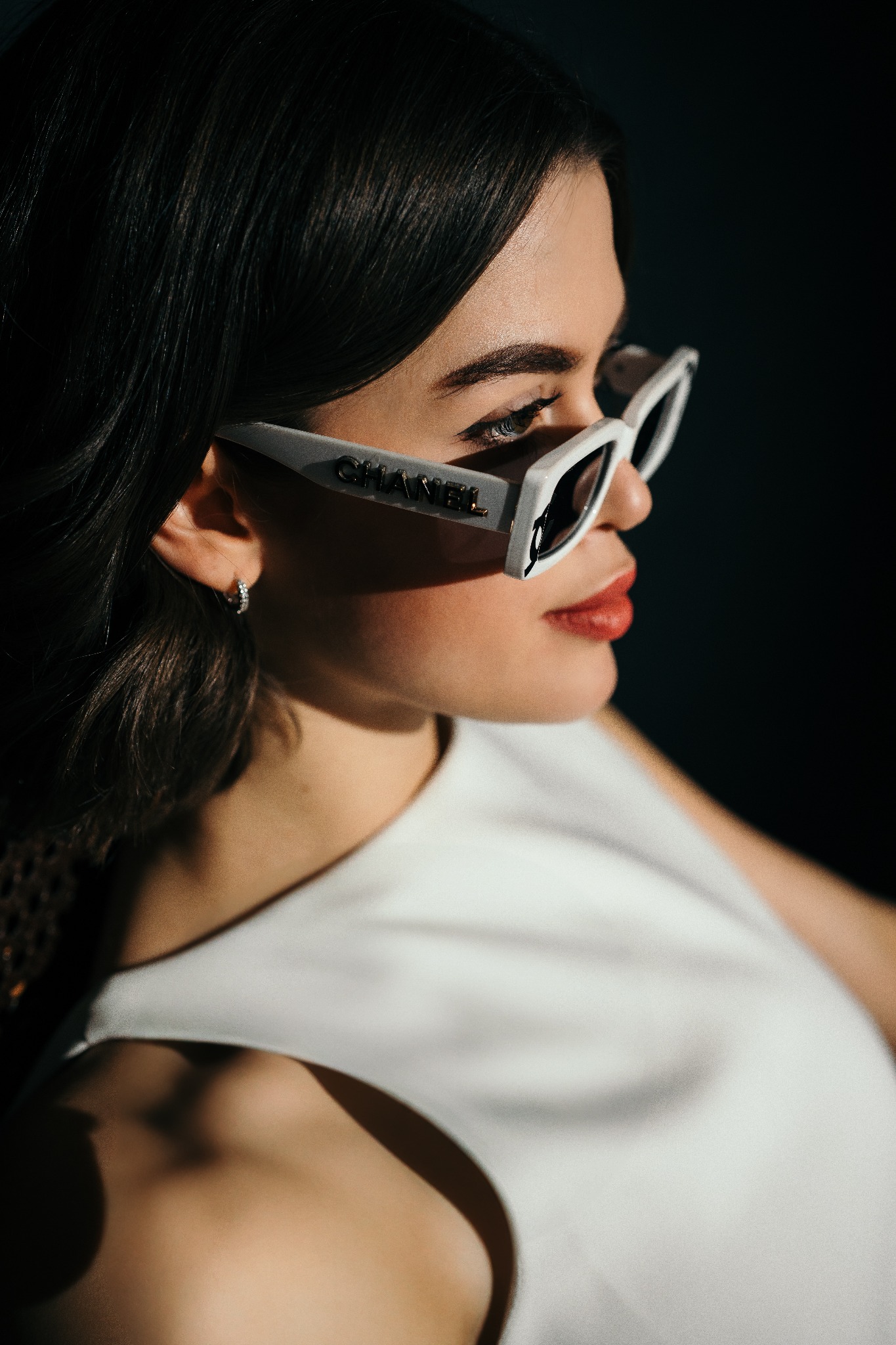 Portret kobiety z profilu w białej sukience i okularach przeciwsłonecznych Chanel, z delikatnym makijażem i kolczykiem w uchu, oświetlona bocznym światłem.