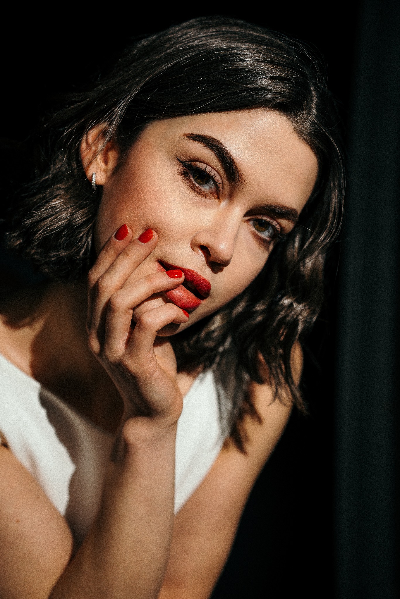 Portret kobiety z krótkimi, ciemnymi włosami, czerwonymi ustami i paznokciami, delikatnie dotykającej twarzy w dramatycznym oświetleniu.