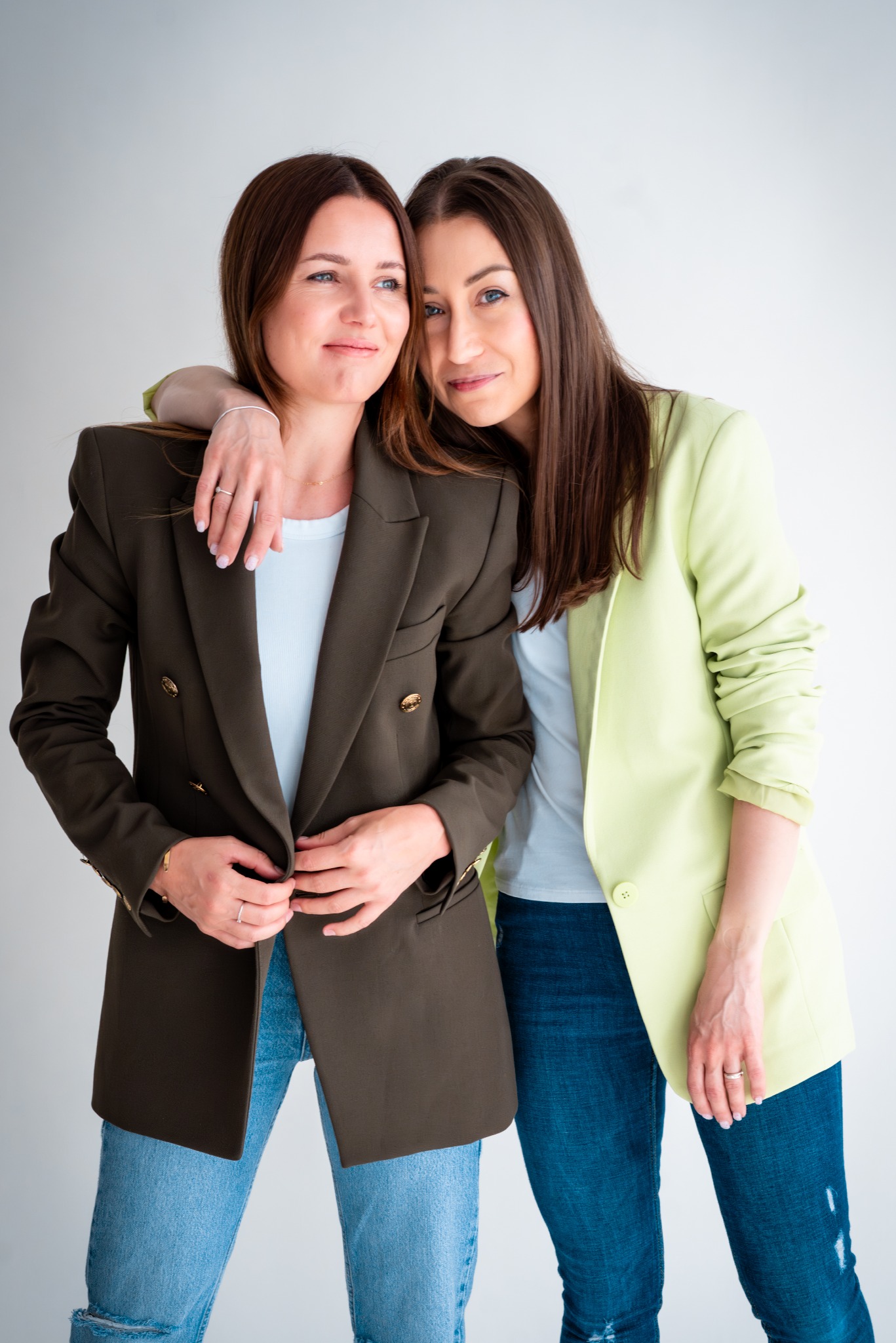 Dwie uśmiechnięte kobiety w marynarkach (khaki i limonka) i dżinsach, pozują na jasnym tle. Jedna obejmuje drugą. Widoczne detale: biżuteria, manicure.