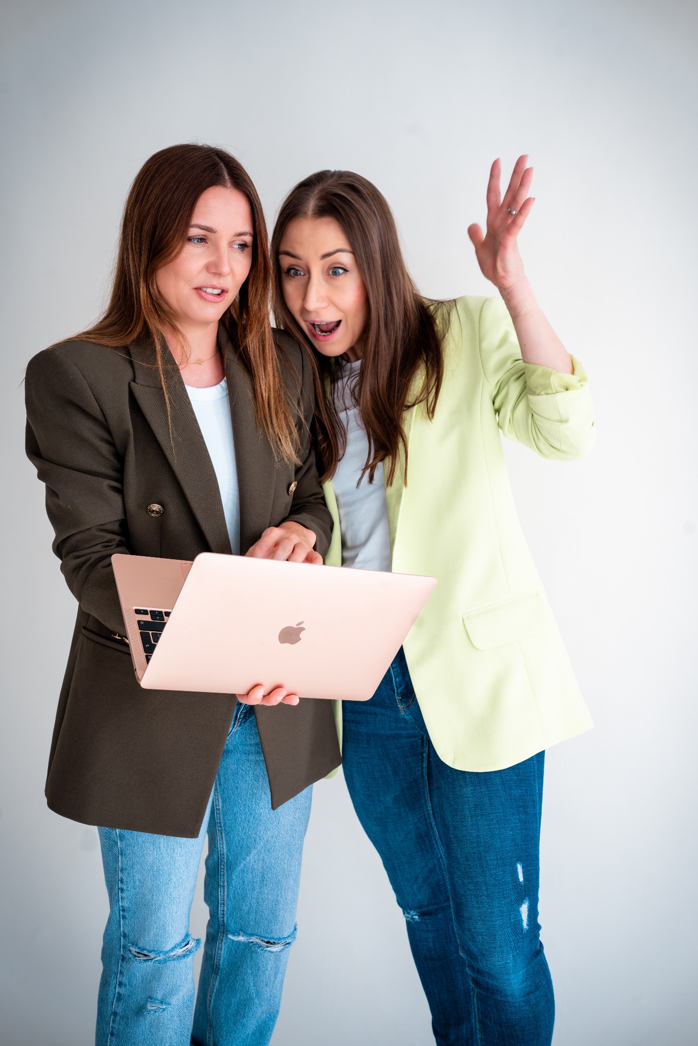 Dwie kobiety w biznesowych stylizacjach, z zaskoczeniem wpatrują się w ekran laptopa w kolorze różowego złota, trzymanego przez jedną z nich na tle białej ściany.