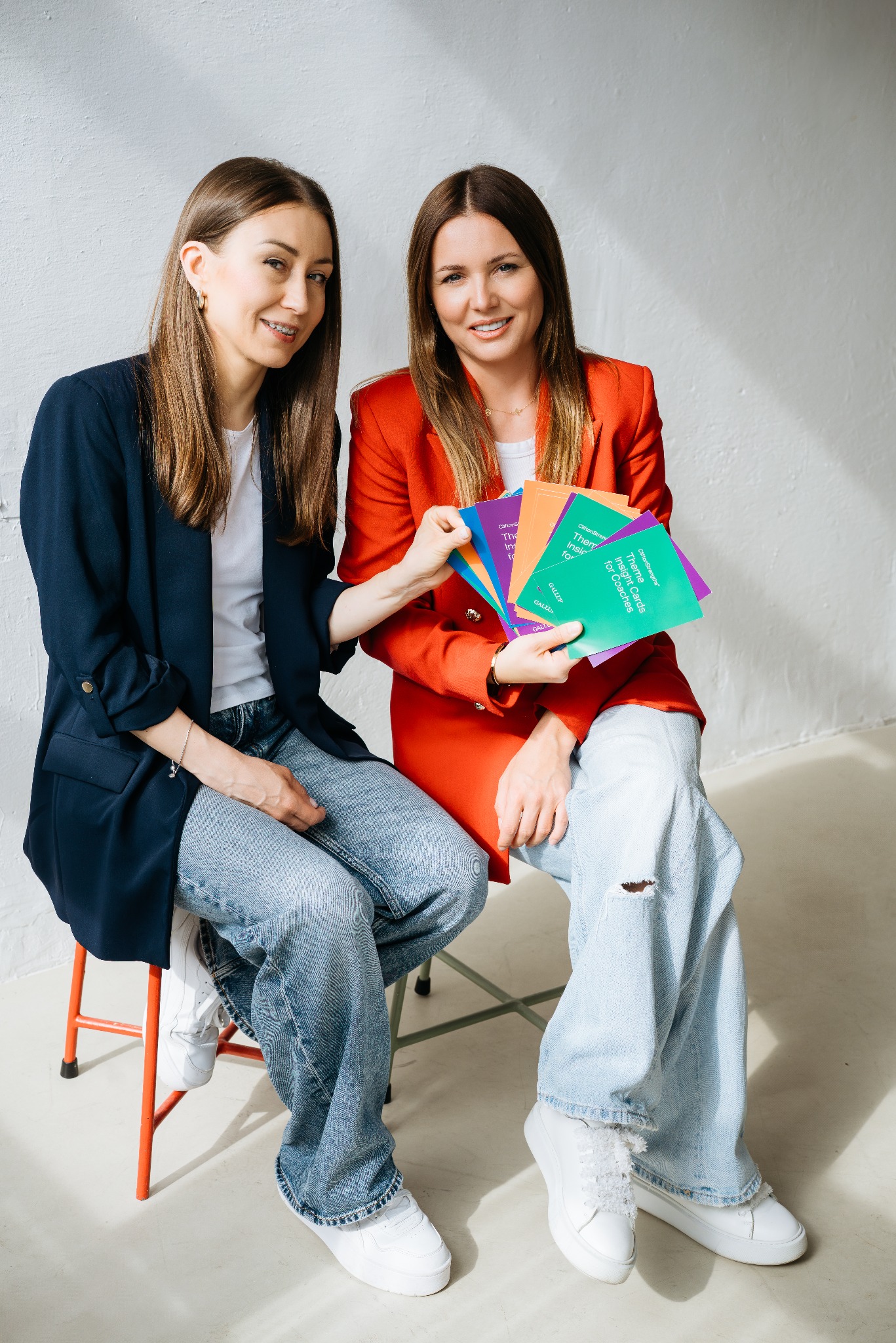 Dwie uśmiechnięte kobiety w stylizowanych ubraniach siedzą obok siebie, prezentując kolorowe karty z napisami na tle białej ściany.