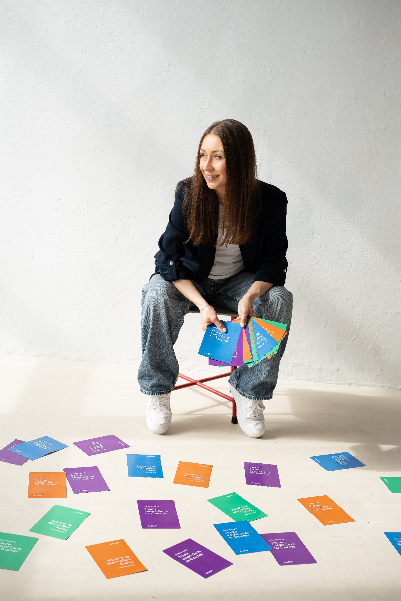 Uśmiechnięta kobieta siedzi na krześle otoczona kolorowymi kartami z napisami, w jasnym pomieszczeniu. Ubrana w jeansy i marynarkę.