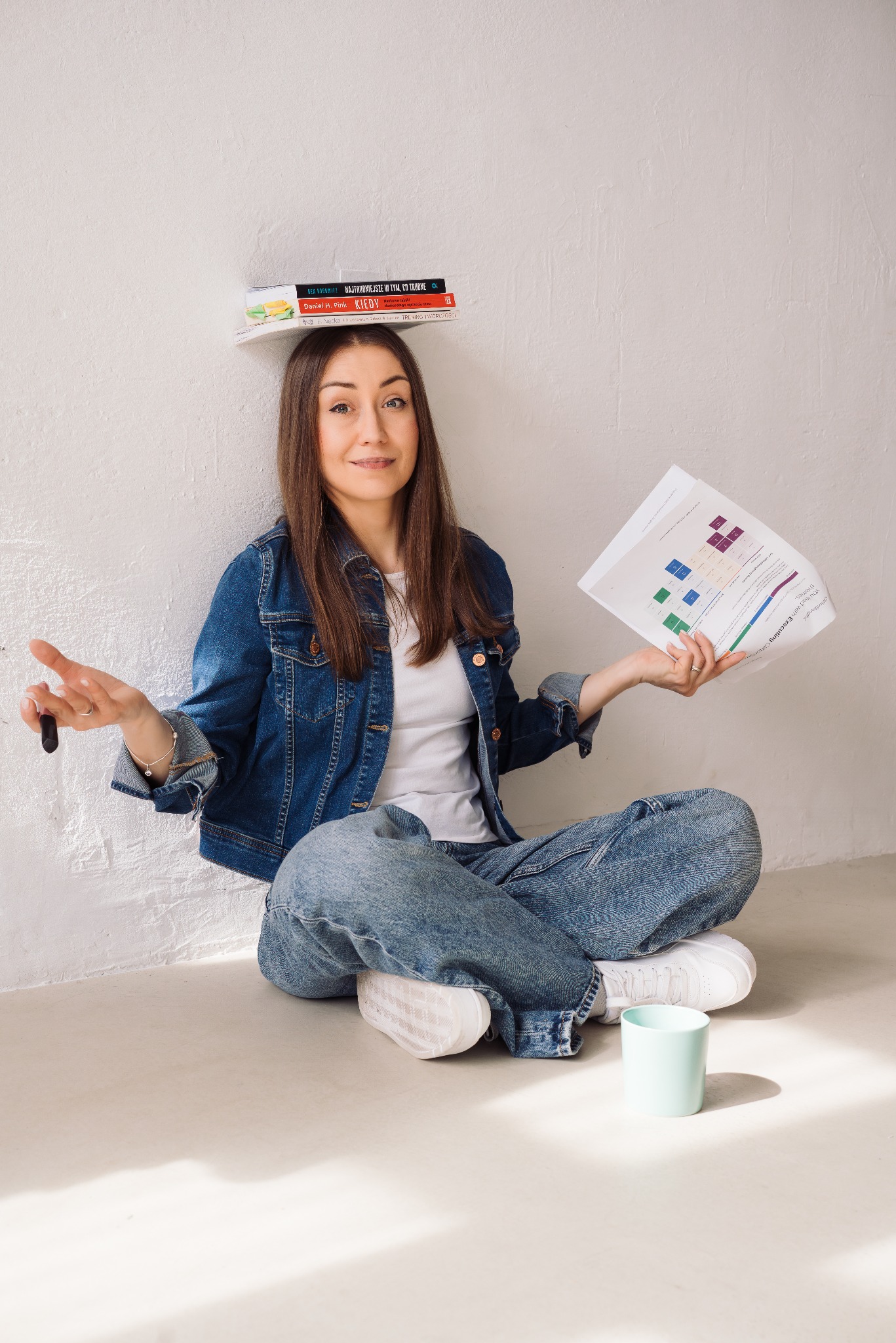 Kobieta siedzi na podłodze z książkami na głowie, trzymając wykres i marker. Uśmiech, dżinsowa kurtka, białe buty. Zabawne ujęcie.