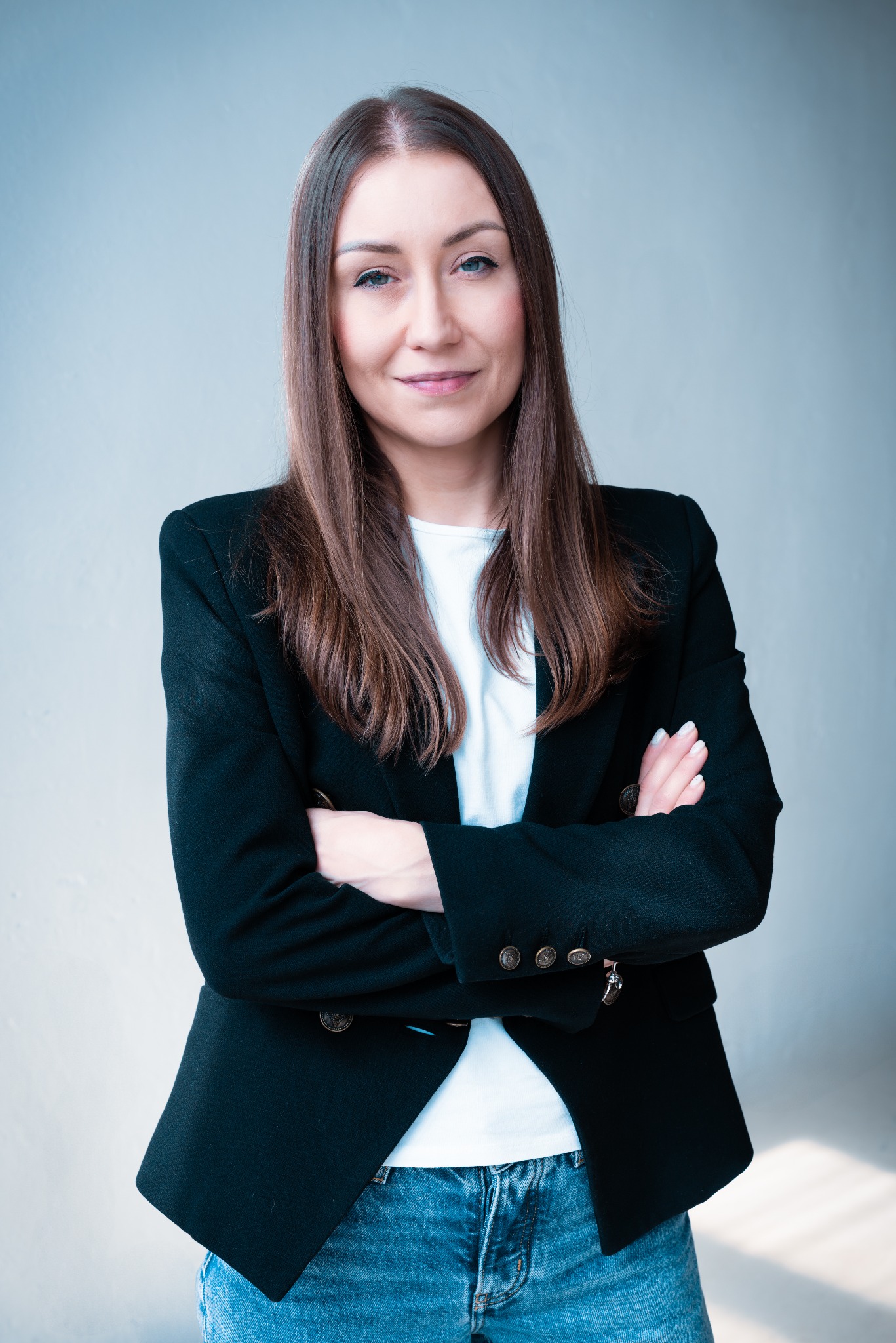 Portret kobiety w czarnej marynarce i jeansach, z założonymi rękami, na jasnym tle. Uśmiech na twarzy, profesjonalny wygląd.