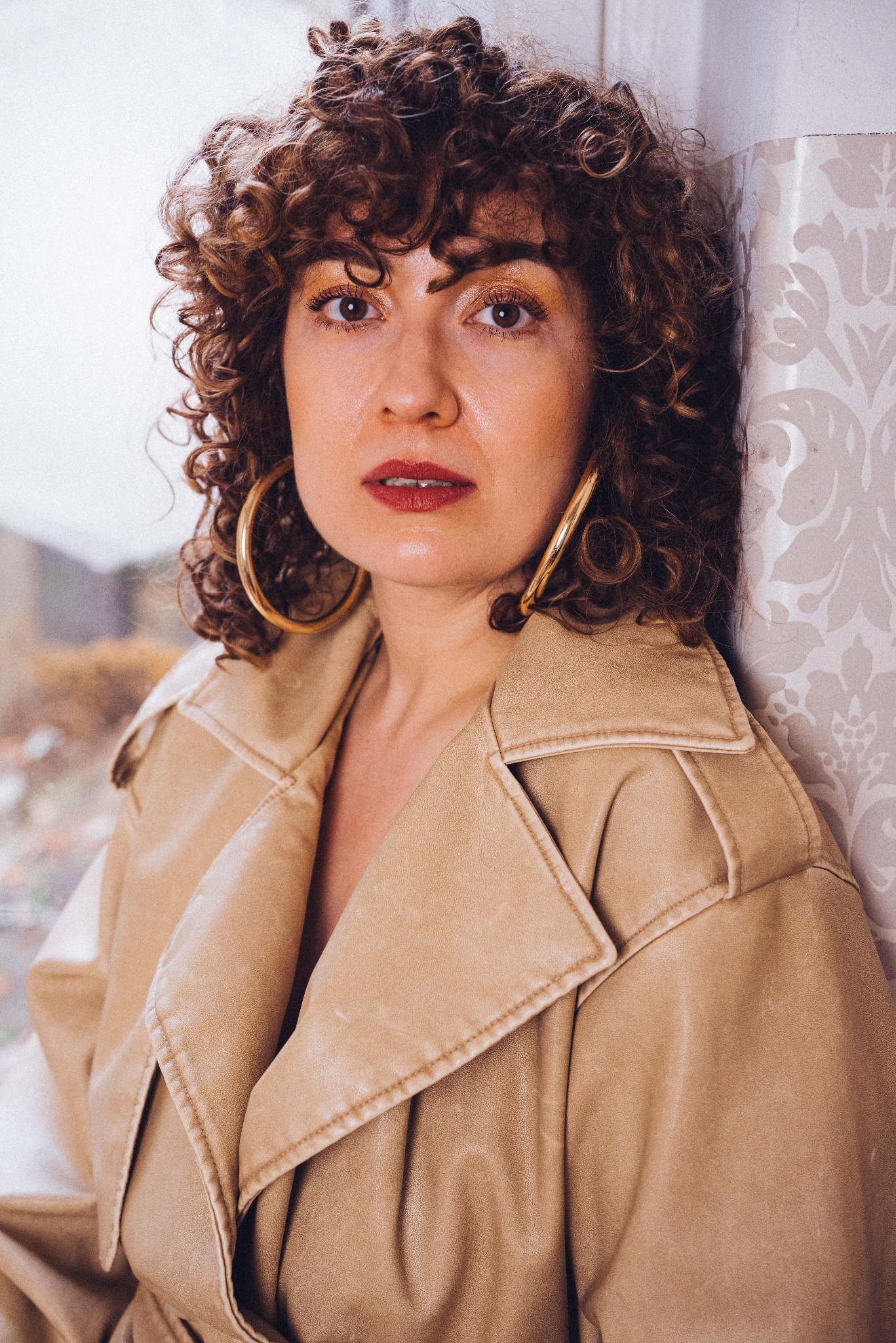 Portret kobiety o kręconych włosach w beżowym płaszczu, z dużymi złotymi kolczykami, oparta o ścianę w kwiatowy wzór, z widocznym fragmentem okna.