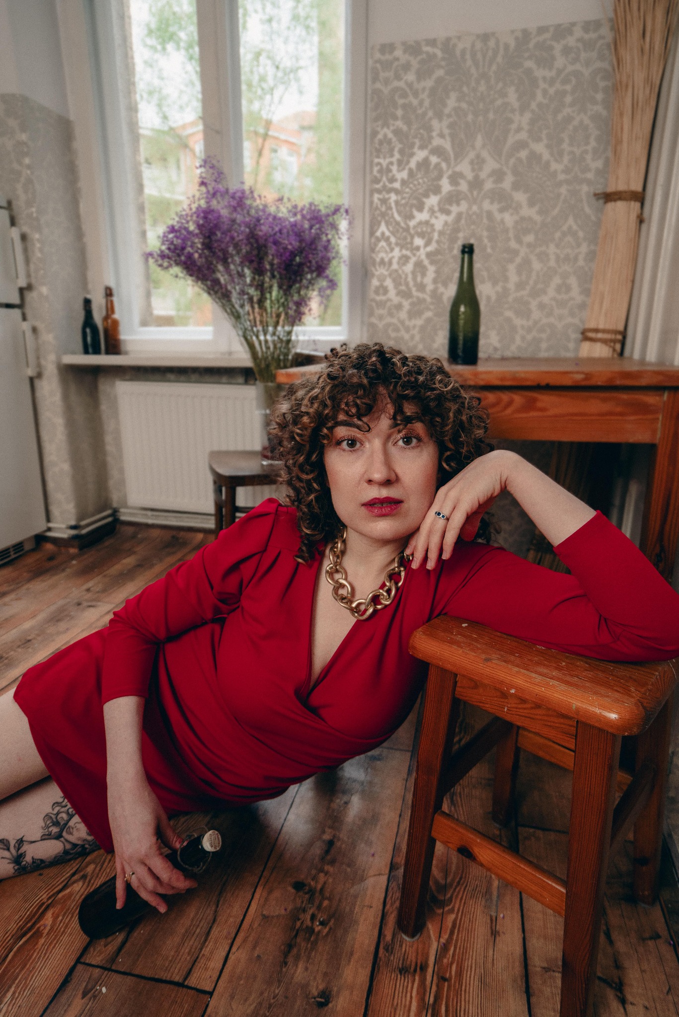 Kobieta w czerwonej sukience, z kręconymi włosami i złotym naszyjnikiem, oparta o drewniany stołek, z butelką w dłoni, w pokoju z kwiatami i wzorzystą tapetą.