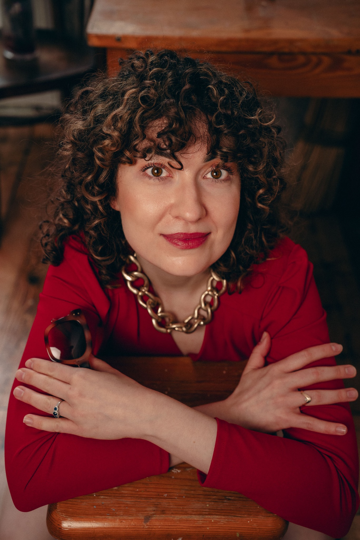 Portret kobiety z kręconymi włosami, w czerwonej sukience i z grubym złotym łańcuchem, spoglądającej w górę, z okularami w dłoni.