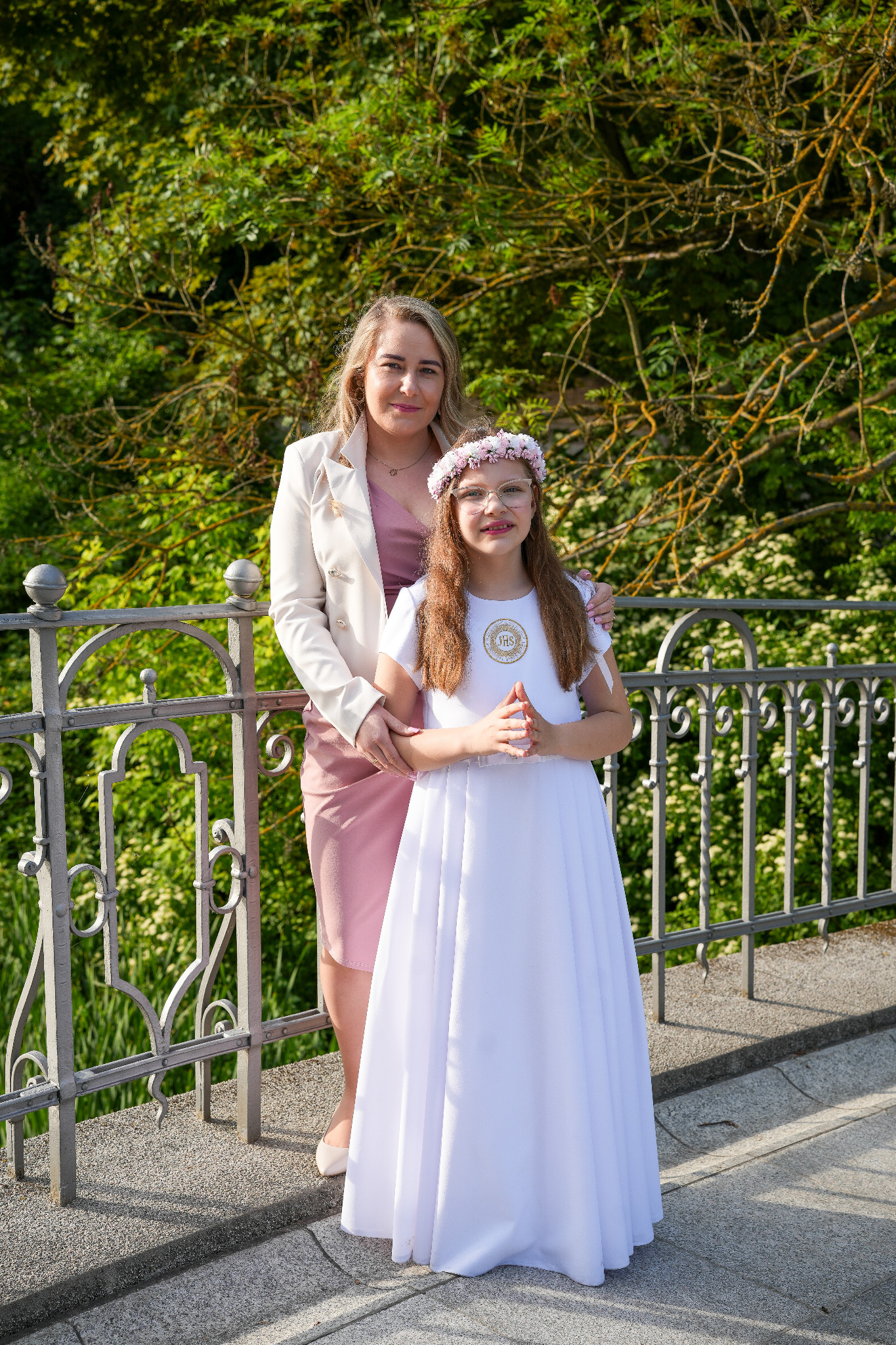 Dziewczynka w białej sukience komunijnej z wiankiem na głowie, pozuje z kobietą w jasnej marynarce na tle zieleni, opierając się o metalową barierkę.