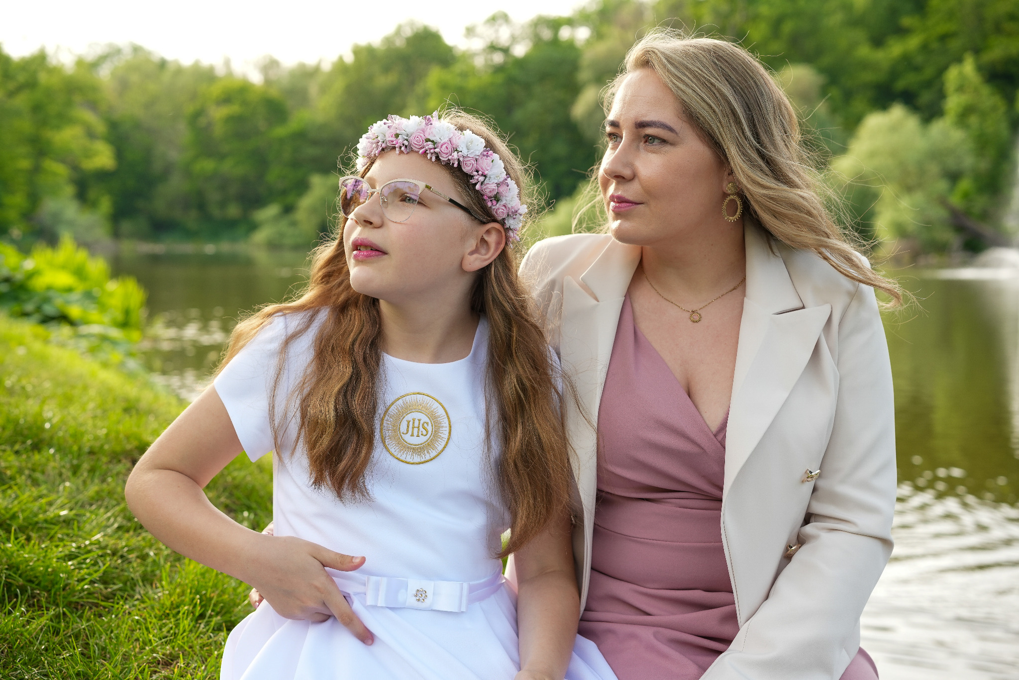 Dziewczynka w okularach i wianku, ubrana w białą sukienkę komunijną, siedzi obok kobiety w różowej sukience i beżowej marynarce na tle jeziora i zieleni.