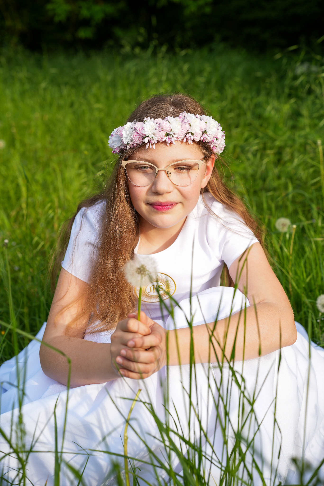 Dziewczynka w okularach i wianku na łące, ubrana w białą sukienkę i koszulkę z symbolem IHS, trzyma dmuchawca. Portret w naturalnym świetle.