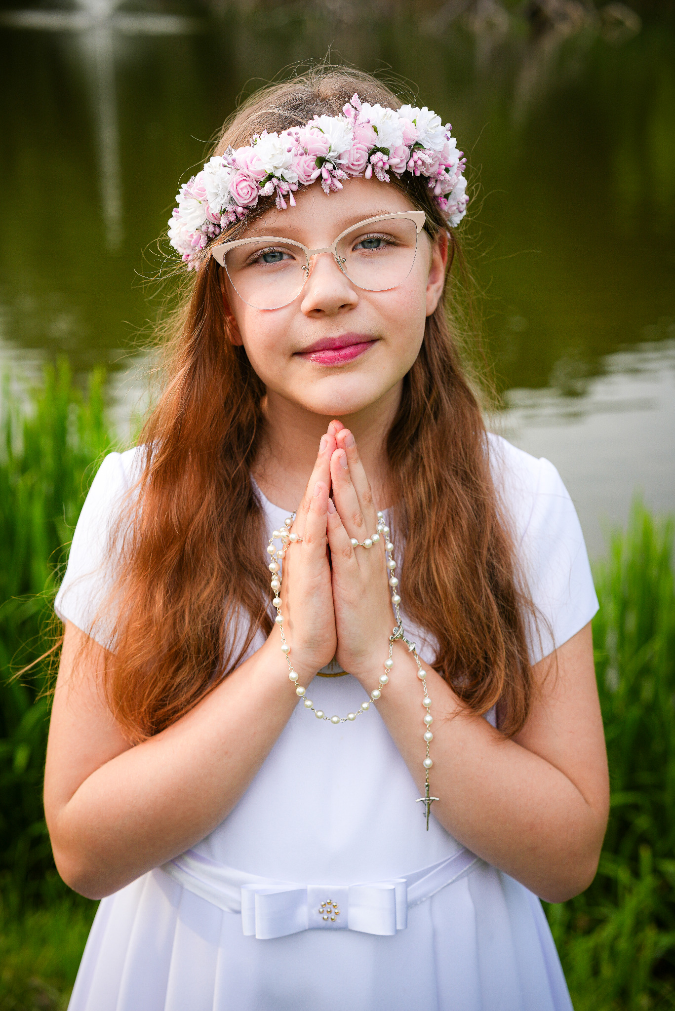Dziewczynka w okularach i wianku z różowych i białych kwiatów, ubrana w białą sukienkę, trzymająca różaniec na tle zieleni i wody.