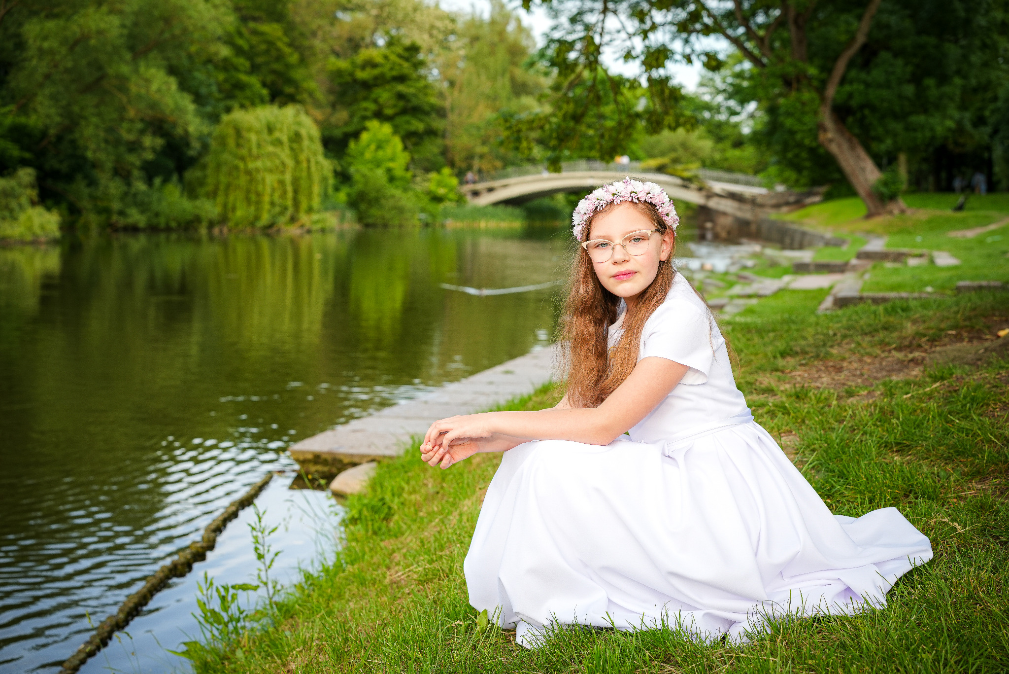 Dziewczynka w białej sukience i wianku siedzi na trawie nad brzegiem jeziora, w tle most i drzewa. Ujęcie z bliska, naturalne światło.
