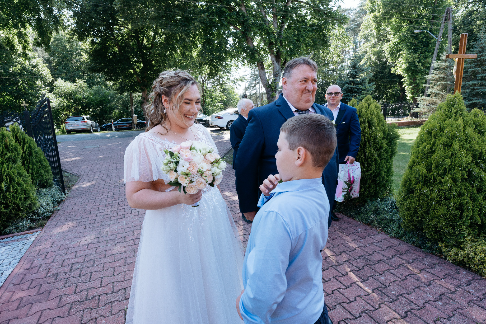 Uśmiechnięta panna młoda w koronkowej sukni trzyma bukiet kwiatów, rozmawiając z chłopcem na tle gości weselnych i zieleni. W tle widoczny krzyż.