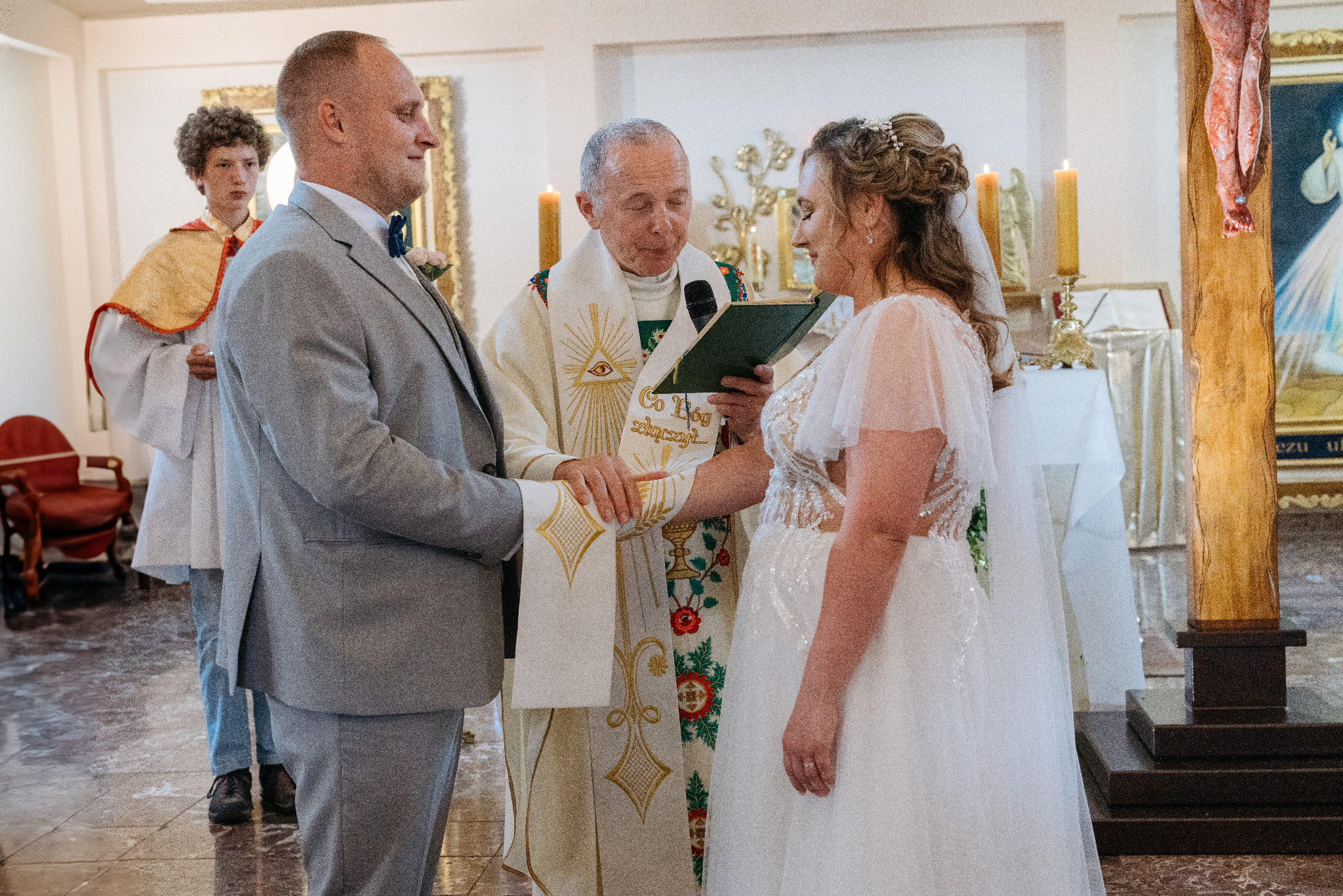 Uroczystość ślubna w kościele: Para młoda podczas ceremonii zaślubin, ksiądz błogosławi dłonie nowożeńców, w tle ministrant i dekoracje kościelne.