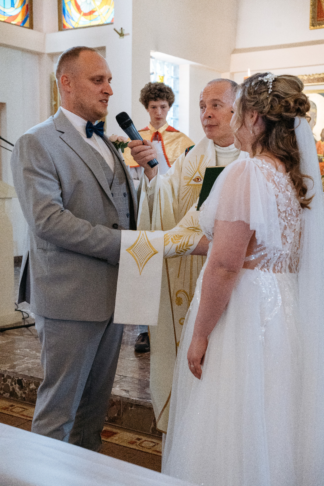 Para młoda podczas ceremonii ślubnej w kościele, ksiądz udziela błogosławieństwa, w tle ministrant. Pan młody w szarym garniturze, panna młoda w białej sukni.