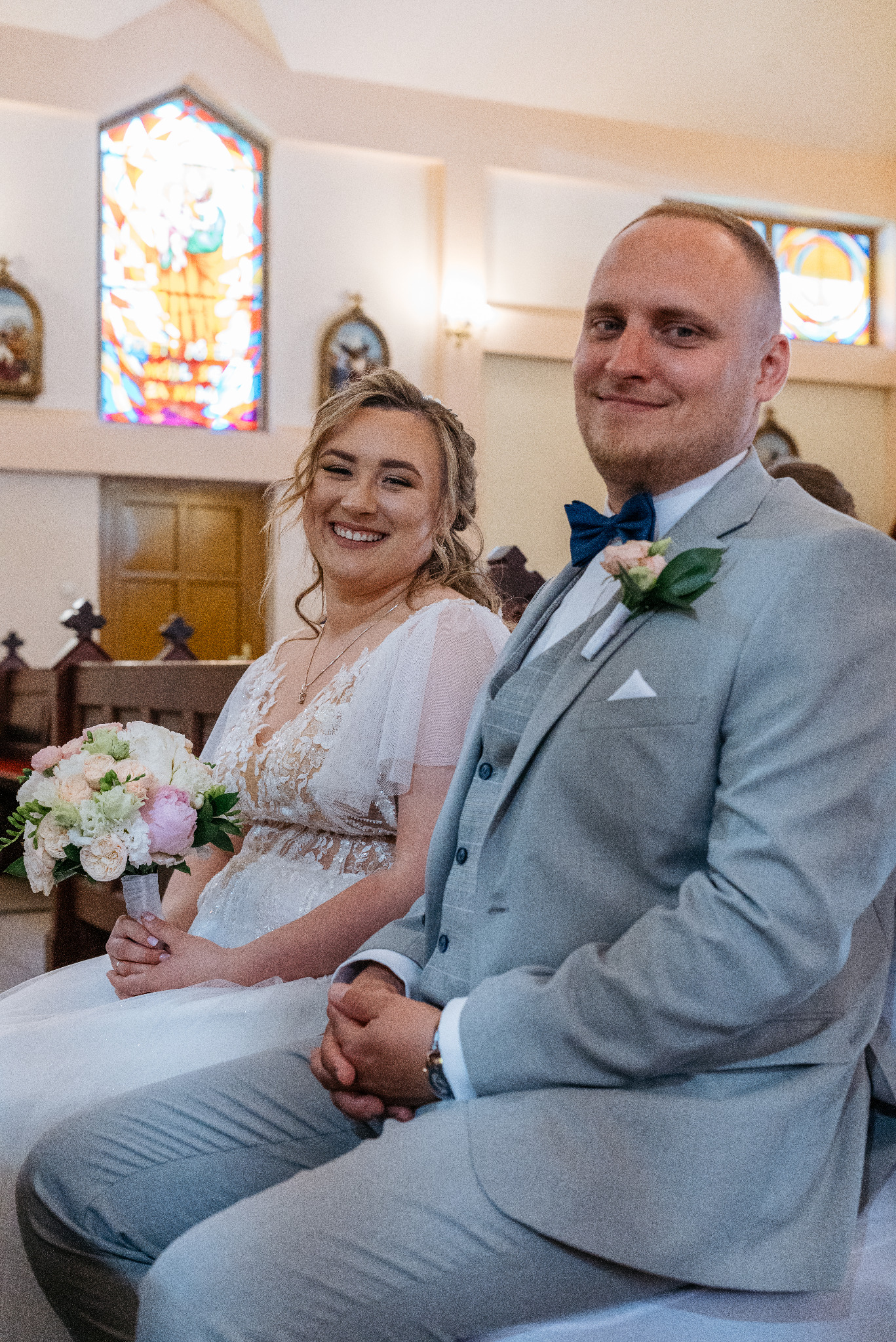 Uśmiechnięta para młoda siedzi w kościele, panna młoda trzyma bukiet, pan młody w szarym garniturze z muszką, w tle witrażowe okno i dekoracje.