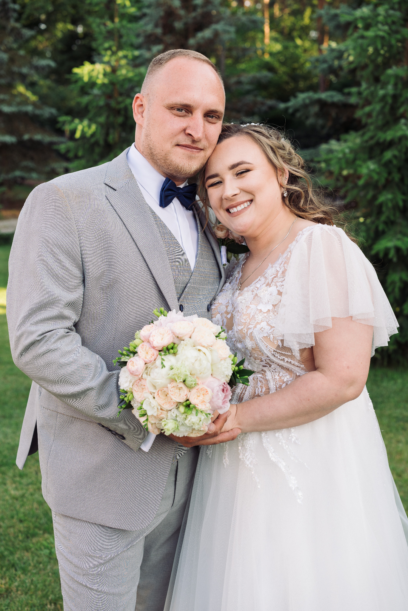 Uśmiechnięta para młoda w objęciach, on w szarym garniturze z muchą, ona w białej sukni z bukietem róż, na tle zieleni.