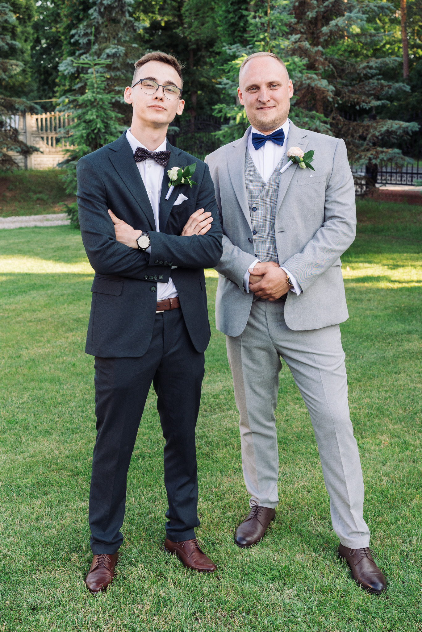 Dwóch eleganckich mężczyzn w garniturach, z muchami i butonierkami, pozuje na trawie przed zielonymi drzewami. Jeden w ciemnym, drugi w szarym garniturze.