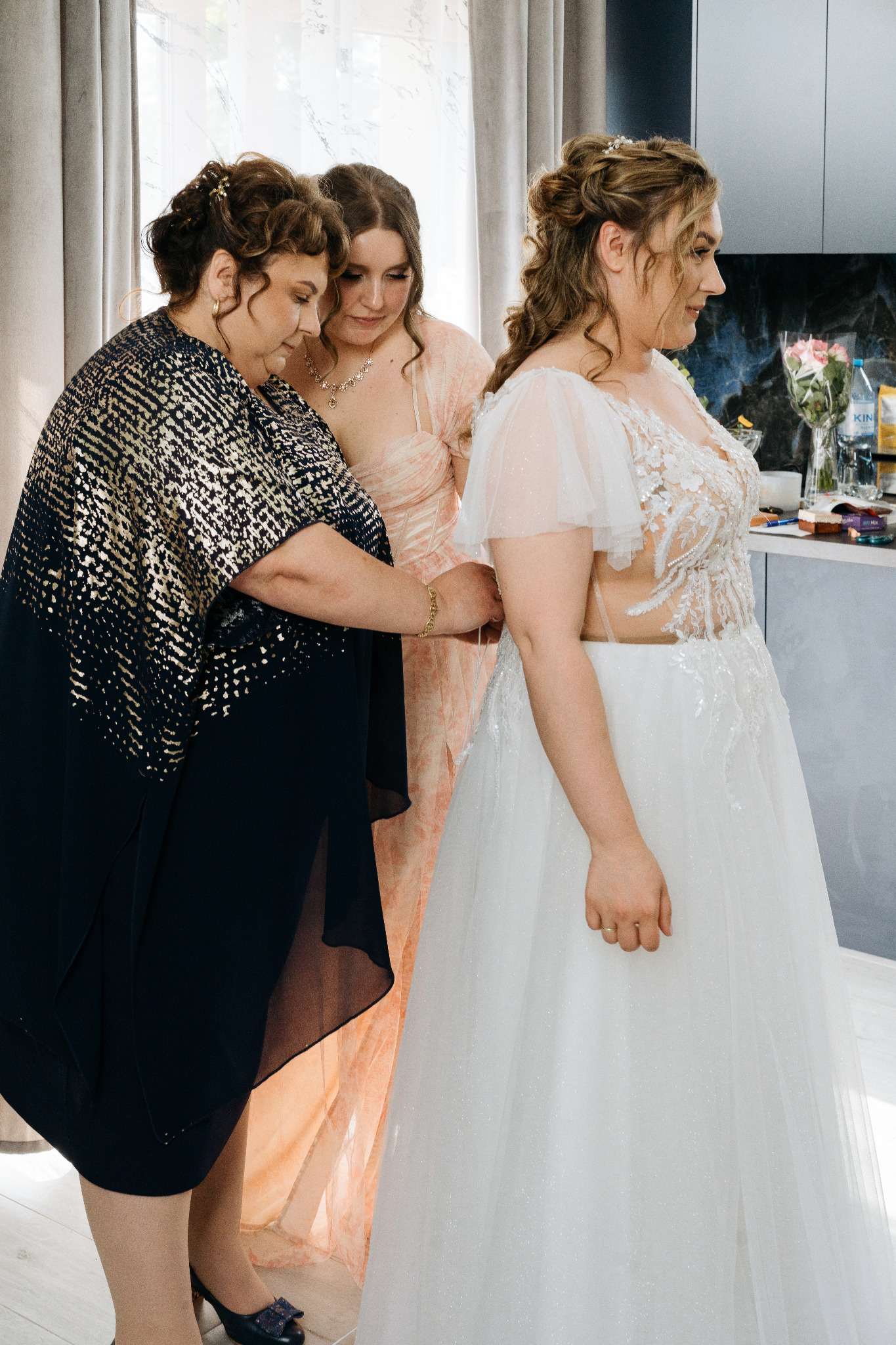 Panna młoda w białej sukni ślubnej, dopinana przez kobietę w ciemnej sukience, obok stoi druhna w jasnej sukni. Przygotowania do ślubu w jasnym wnętrzu.