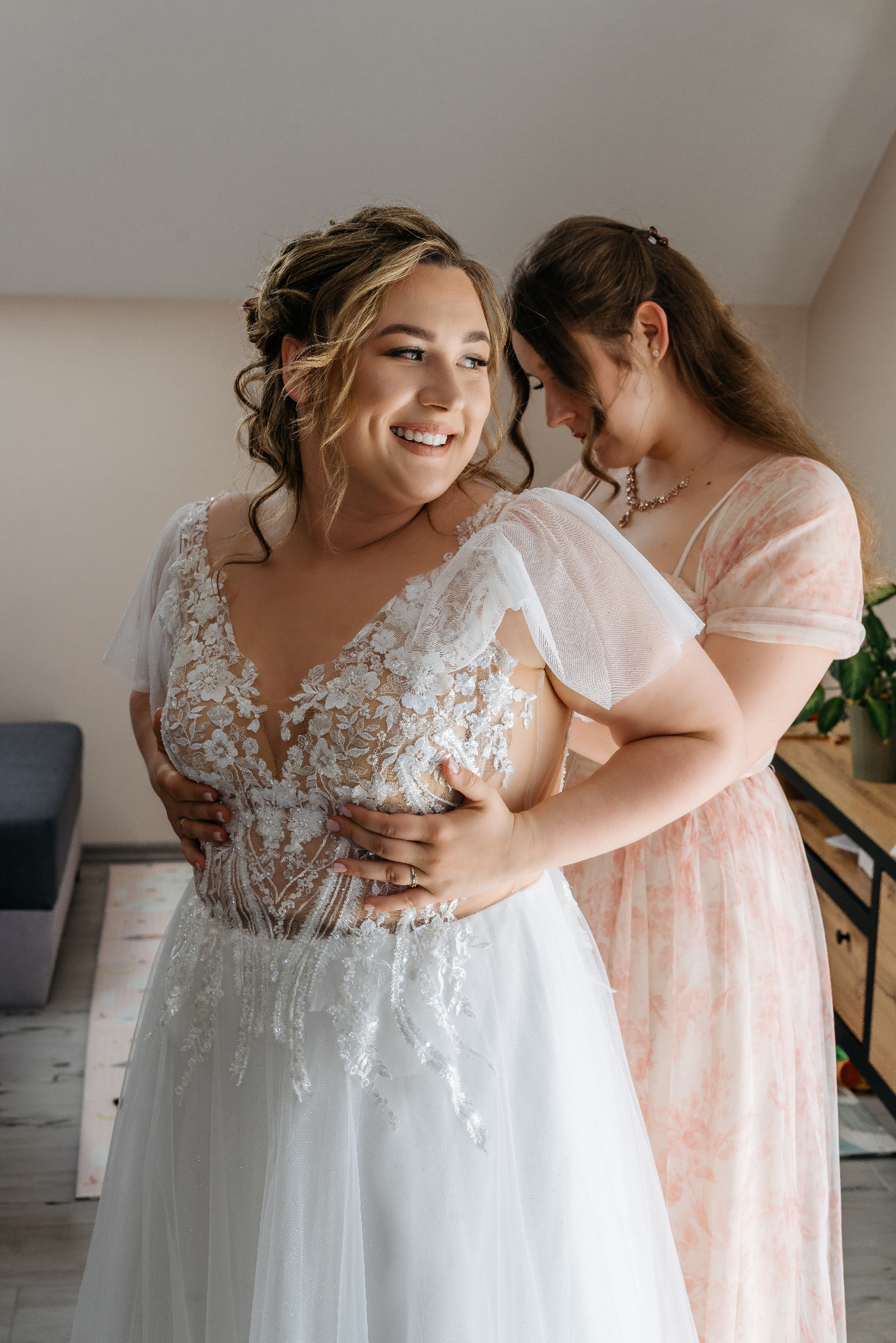 Uśmiechnięta panna młoda w koronkowej sukni ślubnej, dopinana przez druhnę w pastelowej sukience, w jasnym wnętrzu podczas przygotowań do ceremonii.