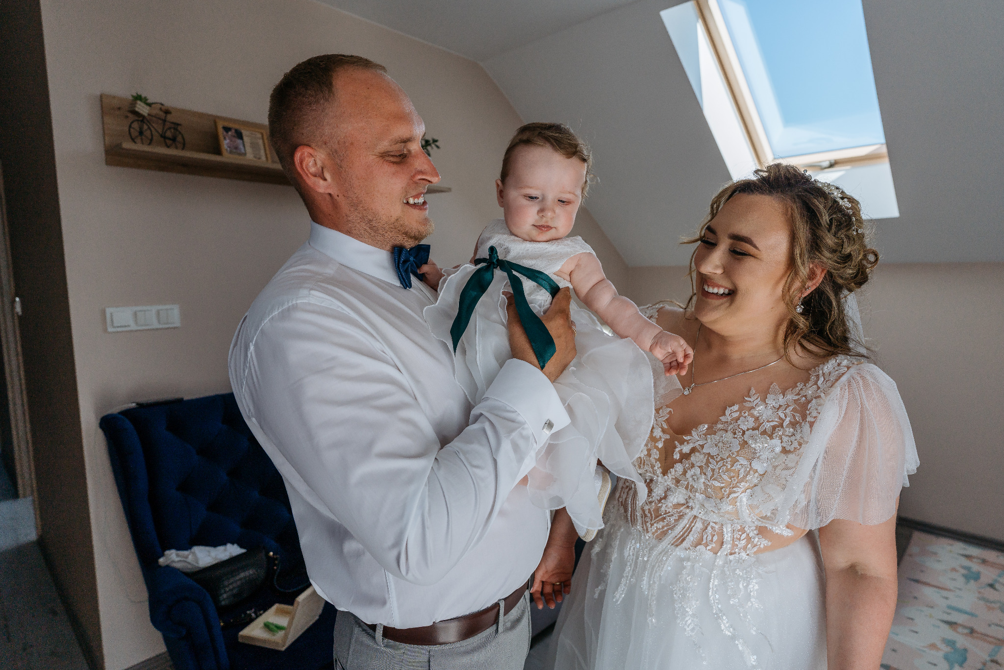 Uśmiechnięta para młoda z niemowlęciem w białej sukience z zieloną wstążką, w jasnym wnętrzu z oknem dachowym. Pan młody w muszce, panna młoda w koronkowej sukni.