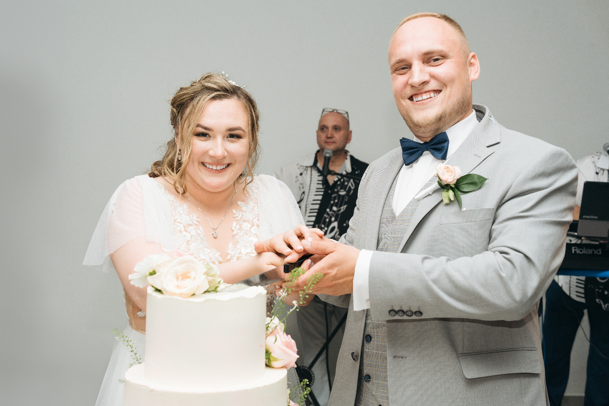 Uśmiechnięta para młoda kroi tort weselny, w tle muzyk z keyboardem. Jasne tło, pastelowe kwiaty na torcie i butonierce pana młodego. Radosna atmosfera.