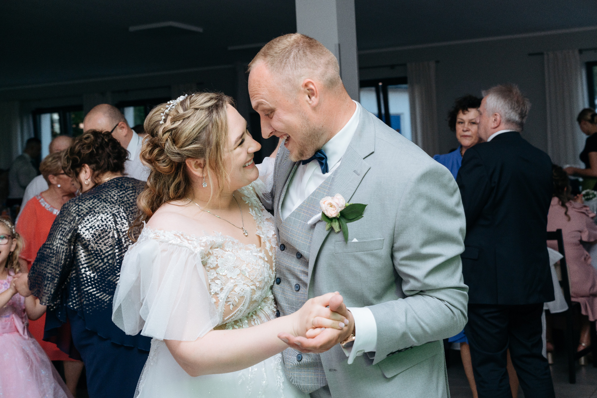 Uśmiechnięta para młoda w tańcu, panna młoda w sukni z koronką, pan młody w szarym garniturze z butonierką, goście w tle, naturalne światło.