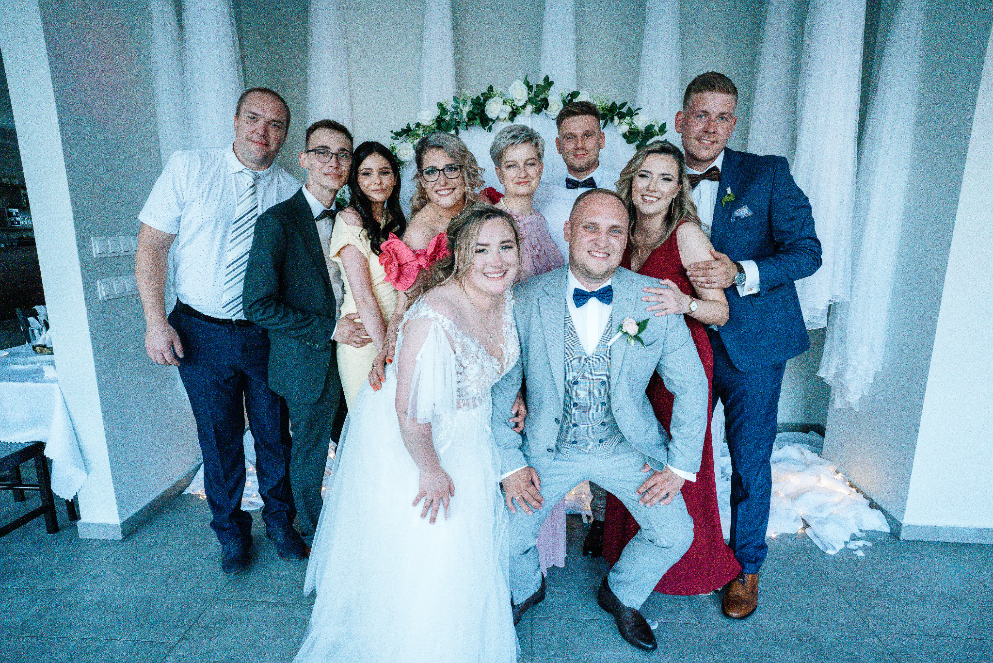 Uśmiechnięta para młoda w otoczeniu gości weselnych pozuje do pamiątkowego zdjęcia na tle dekoracji z białych kwiatów i delikatnych światełek.