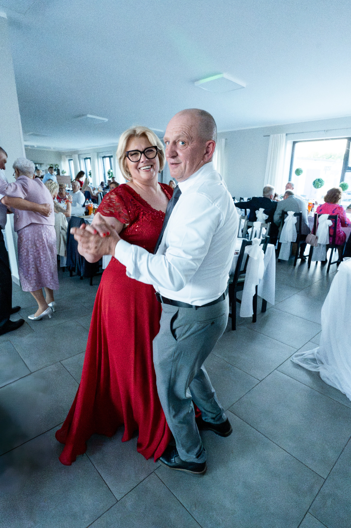 Uśmiechnięta para w tańcu na sali balowej, kobieta w czerwonej sukni, mężczyzna w białej koszuli i szarych spodni, goście w tle, dekoracje stołów.