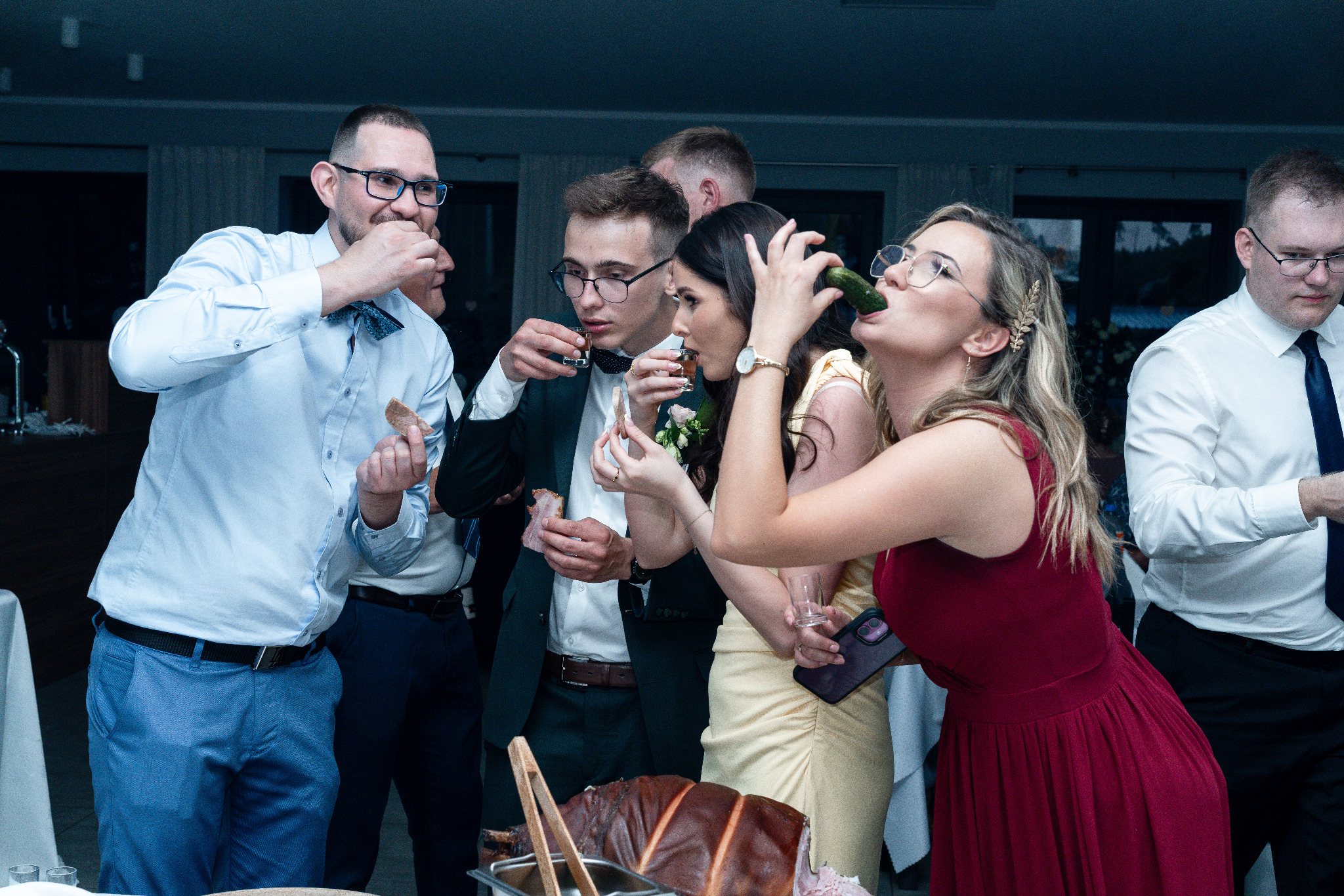 Grupa gości weselnych w trakcie toastu: panowie z kieliszkami, pani pije ogórka kiszonego. Na pierwszym planie prosiak pieczony. Uczestnicy w eleganckich strojach.