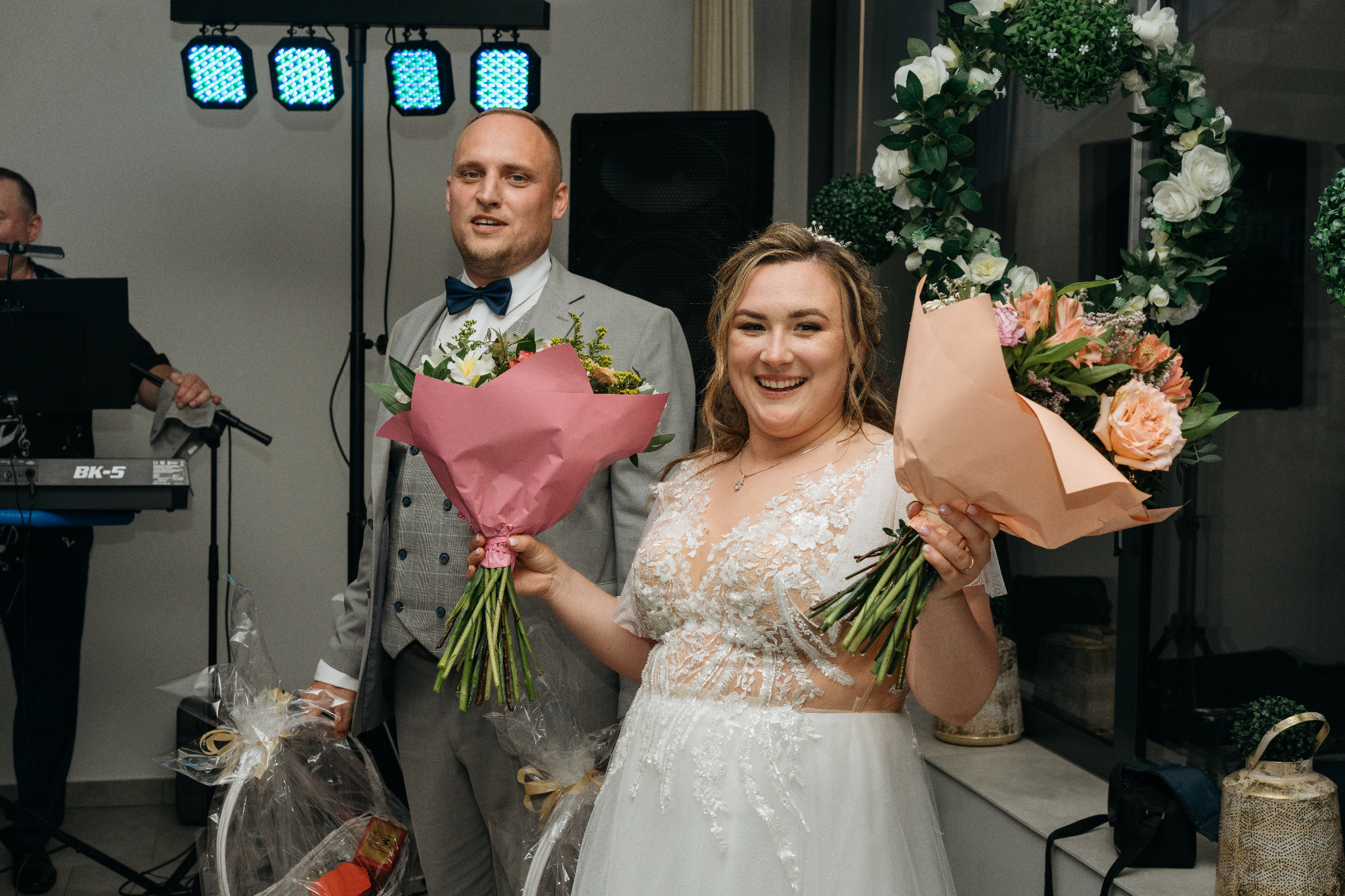 Uśmiechnięta para młoda z bukietami kwiatów, na tle dekoracji weselnych i muzyka na żywo. Panna młoda w koronkowej sukni, pan młody w szarej marynarce.