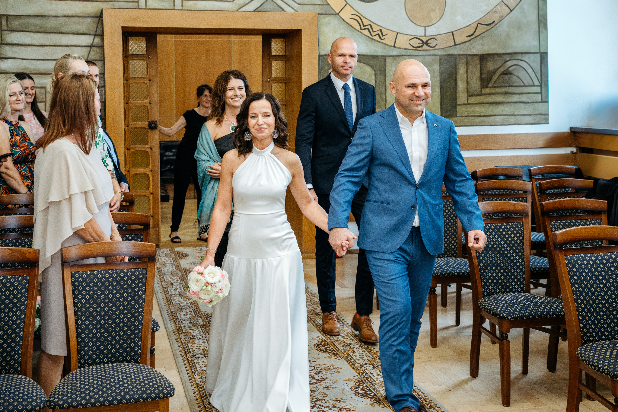 Para młoda, trzymając się za ręce, wchodzi do sali ślubów, za nimi goście. Panna młoda w białej sukni, pan młody w niebieskim garniturze. Uśmiechnięci, w jasnym wnętrzu.