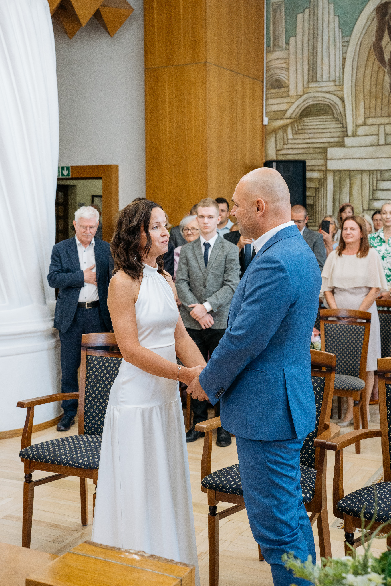 Para młoda trzymająca się za ręce podczas ceremonii ślubnej w jasnym wnętrzu z gośćmi w tle i dekoracyjnym malowidłem na ścianie.