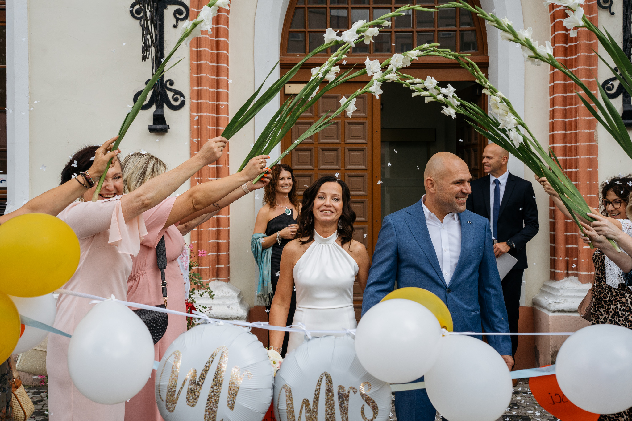 Para młoda przechodzi pod szpalerem z kwiatów i balonów w Szczecinie. Uśmiechnięci nowożeńcy w eleganckich strojach, w tle goście weselni i budynek z ceglaną fasadą.