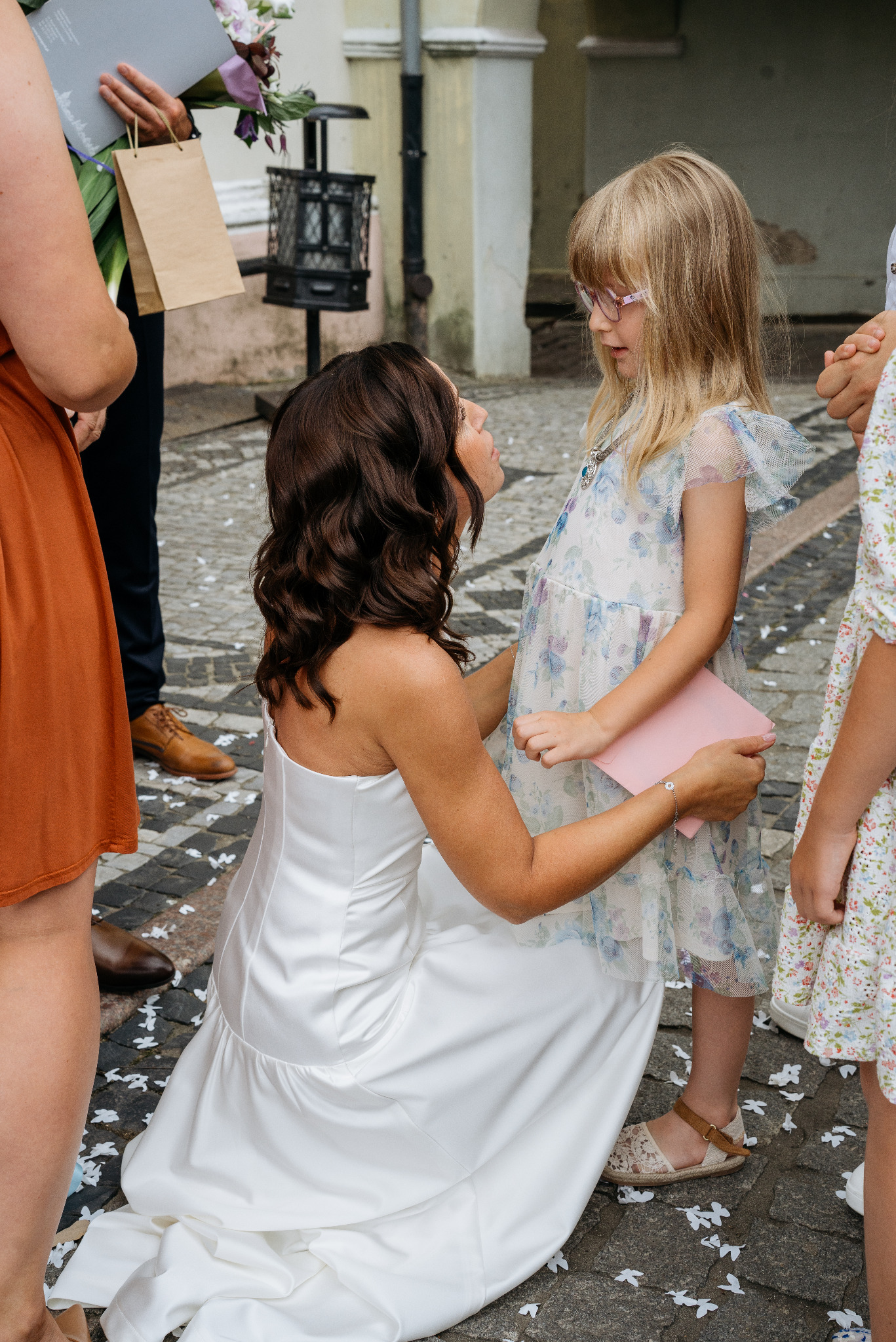 Panna młoda w białej sukni klęczy, patrząc na dziewczynkę w okularach i kwiecistej sukience, trzymającą różową kopertę. W tle goście weselni i bruk z rozsypanymi płatkami.