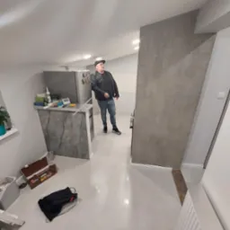 Wnętrze po remoncie: gładkie, jasne ściany, biała podłoga z połyskiem, szara ściana dekoracyjna z fakturą, mężczyzna w czarnej kurtce ogląda sufit, widoczne fragmenty kuchni.