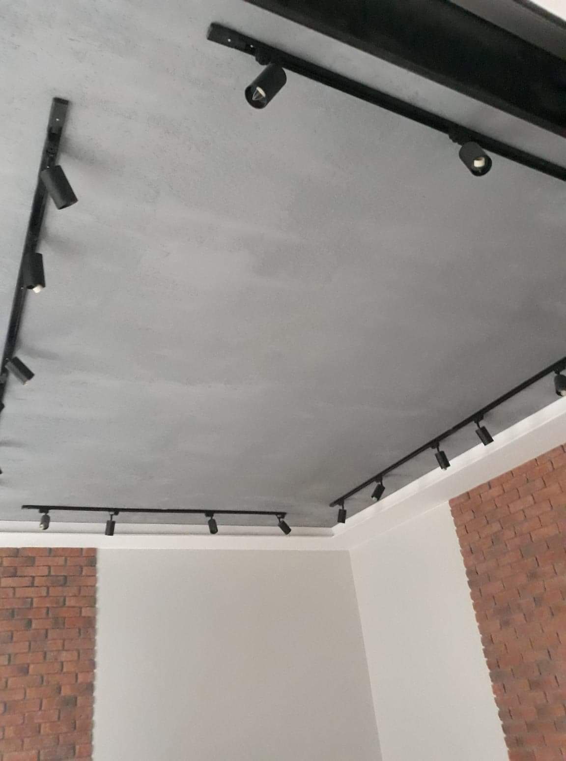 Szary sufit z zamontowanym systemem szynowym z czarnymi reflektorami punktowymi, ściany częściowo wykończone cegłą dekoracyjną.