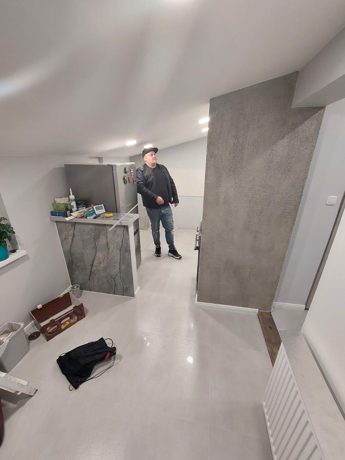 Wnętrze po remoncie: gładkie, jasne ściany, biała podłoga z połyskiem, szara ściana dekoracyjna z fakturą, mężczyzna w czarnej kurtce ogląda sufit, widoczne fragmenty kuchni.