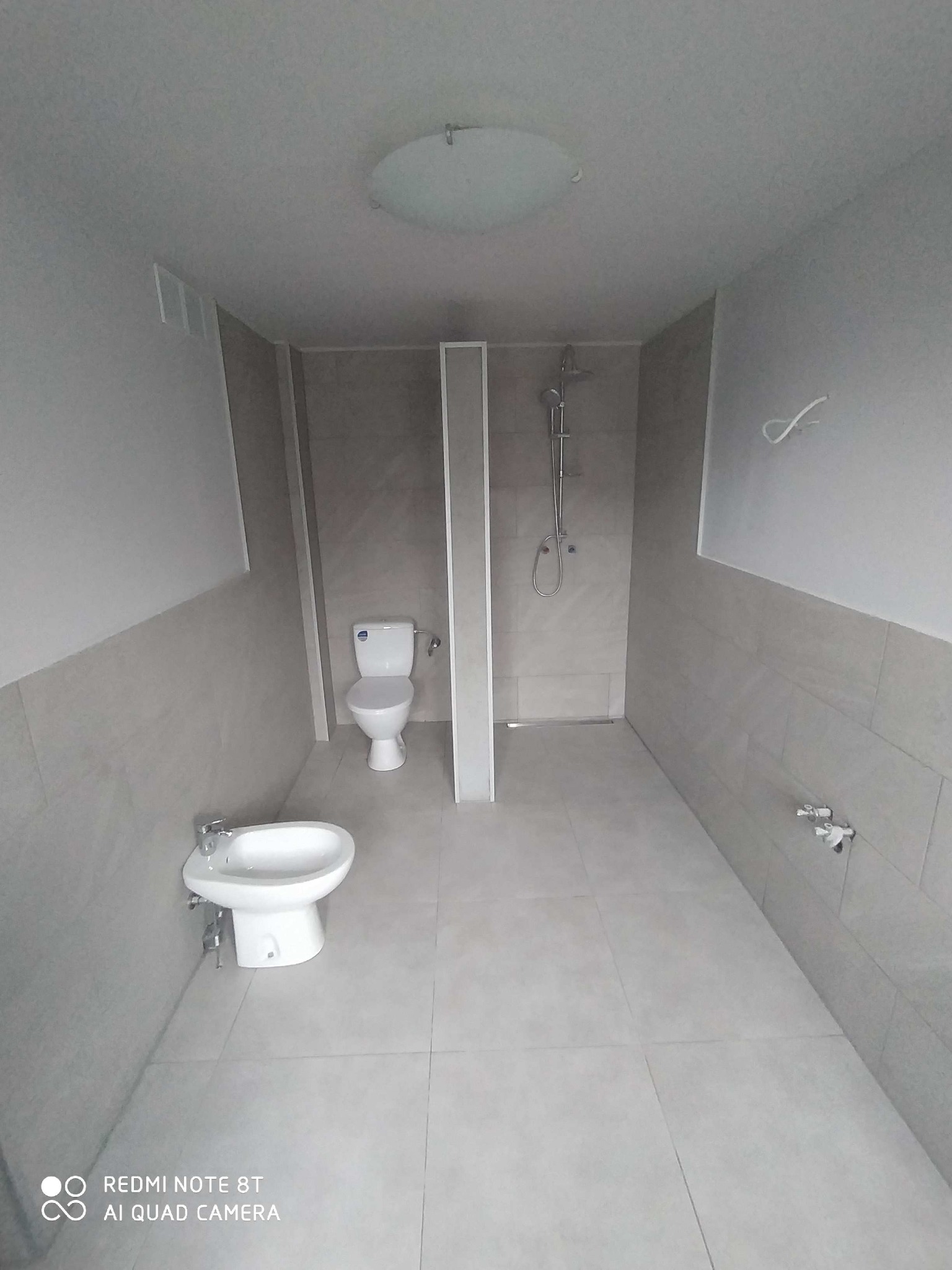 Wykończona łazienka z szarymi płytkami, białą toaletą, bidetem i kabiną prysznicową typu walk-in z chromowaną baterią, widoczne wystające kable elektryczne.