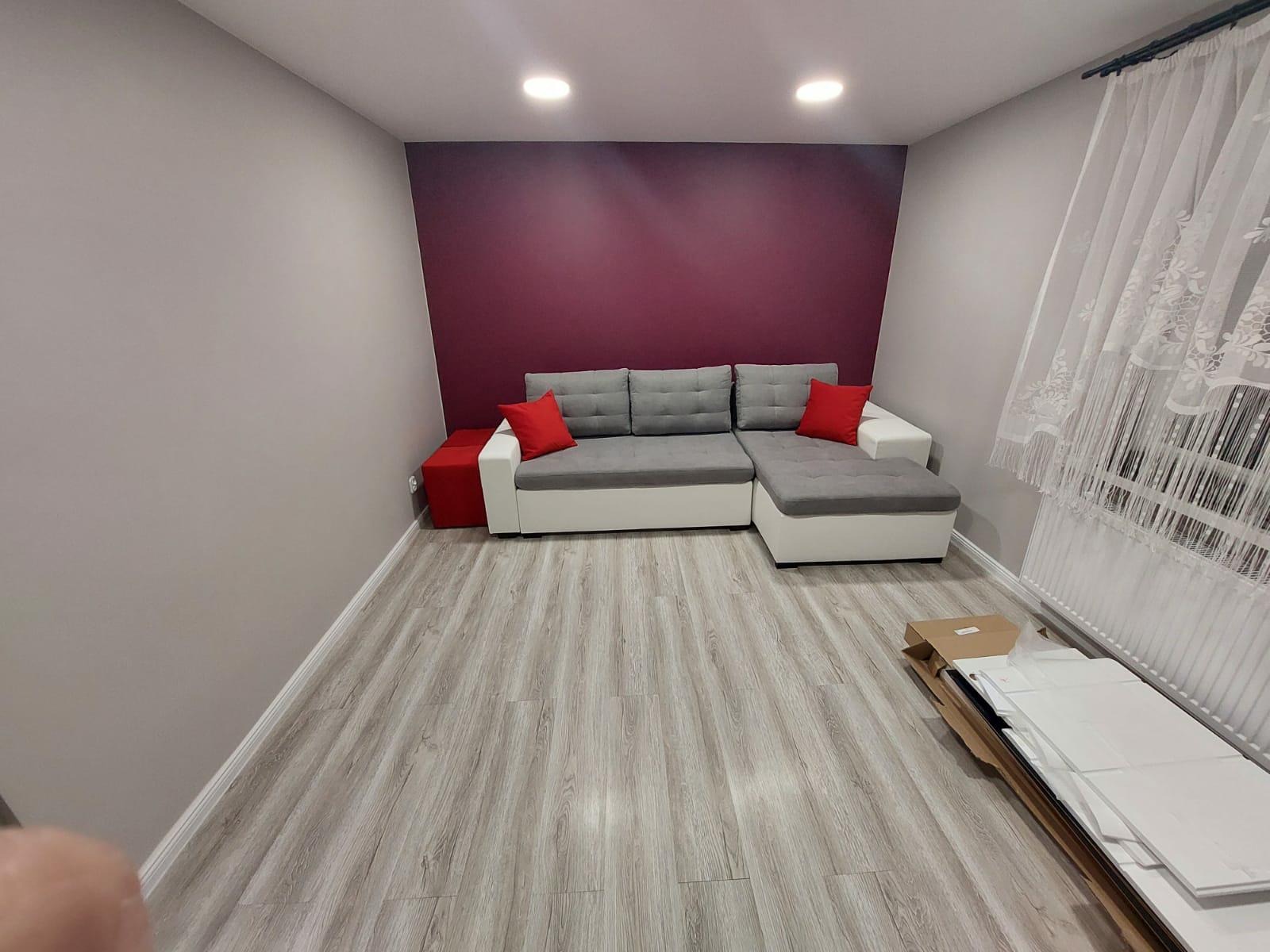 Szary narożnik z czerwonymi poduszkami na tle bordowej ściany, jasna podłoga imitująca drewno, białe zasłony i rozpakowane panele podłogowe w rogu pokoju.