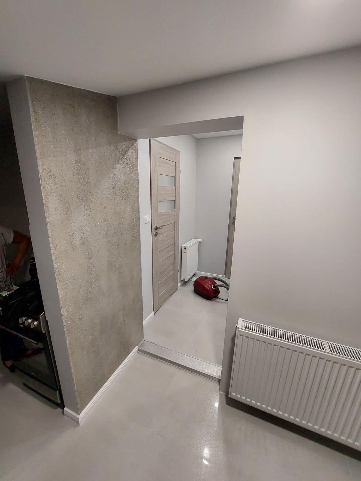 Wnętrze po remoncie: szara ściana z efektem dekoracyjnym, gładka podłoga, biały grzejnik, drzwi z przeszkleniami i czerwony odkurzacz w tle.