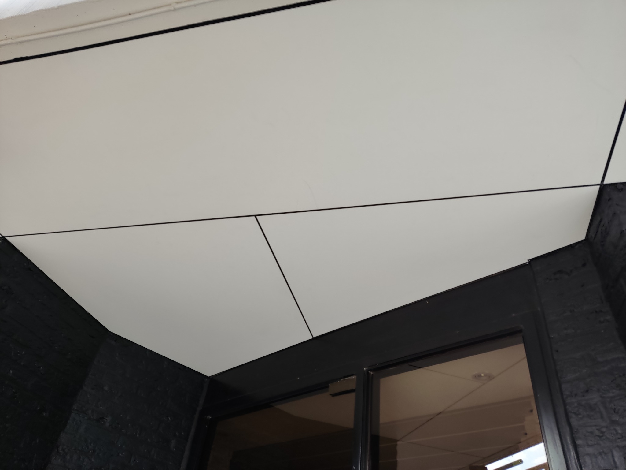 Nowoczesny sufit z białych paneli, z czarnymi liniami podziału, widok z dołu, z fragmentem czarnej ściany i ramy drzwi.